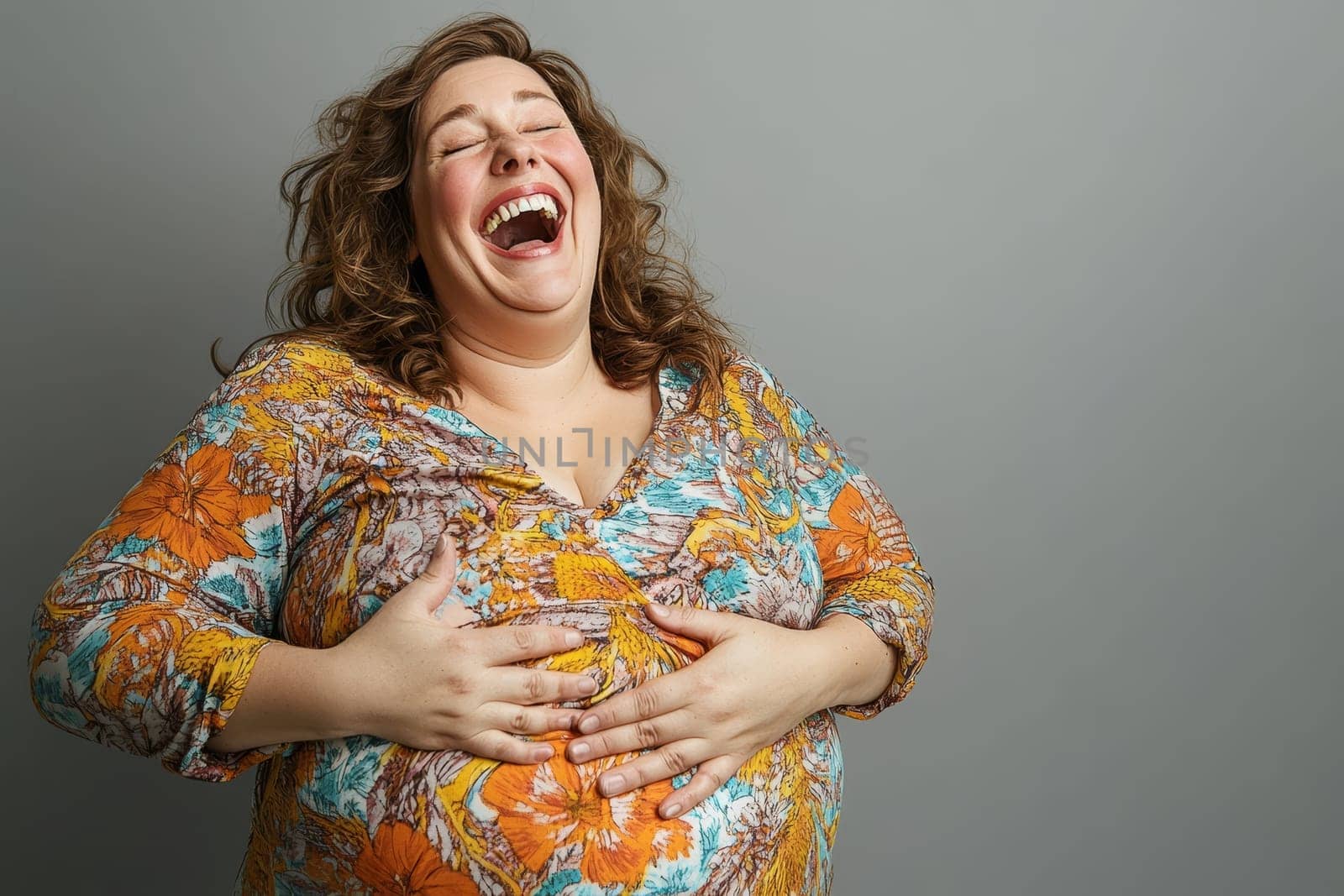 Curvy woman with curly hair wearing colorful floral top laughing wholeheartedly while holding her belly, embodying joy and body positivity against neutral backdrop.