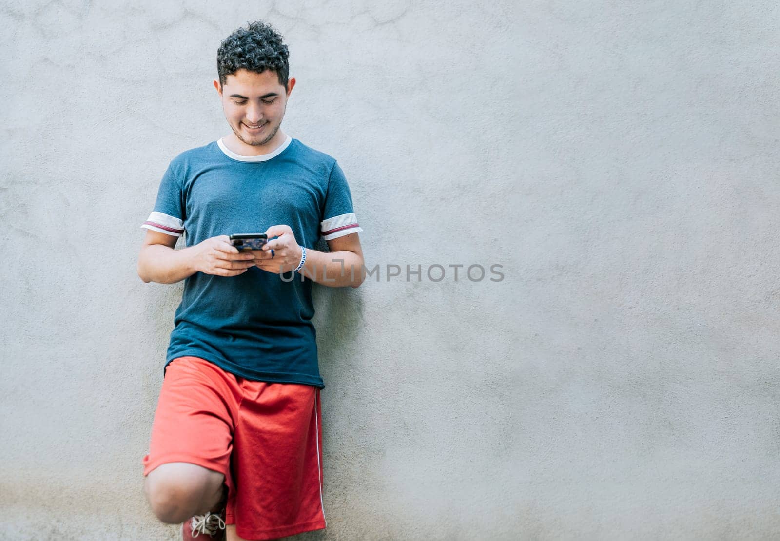Happy guy leaning on a wall using smartphone. Smiling young man leaning on a wall sending a text message