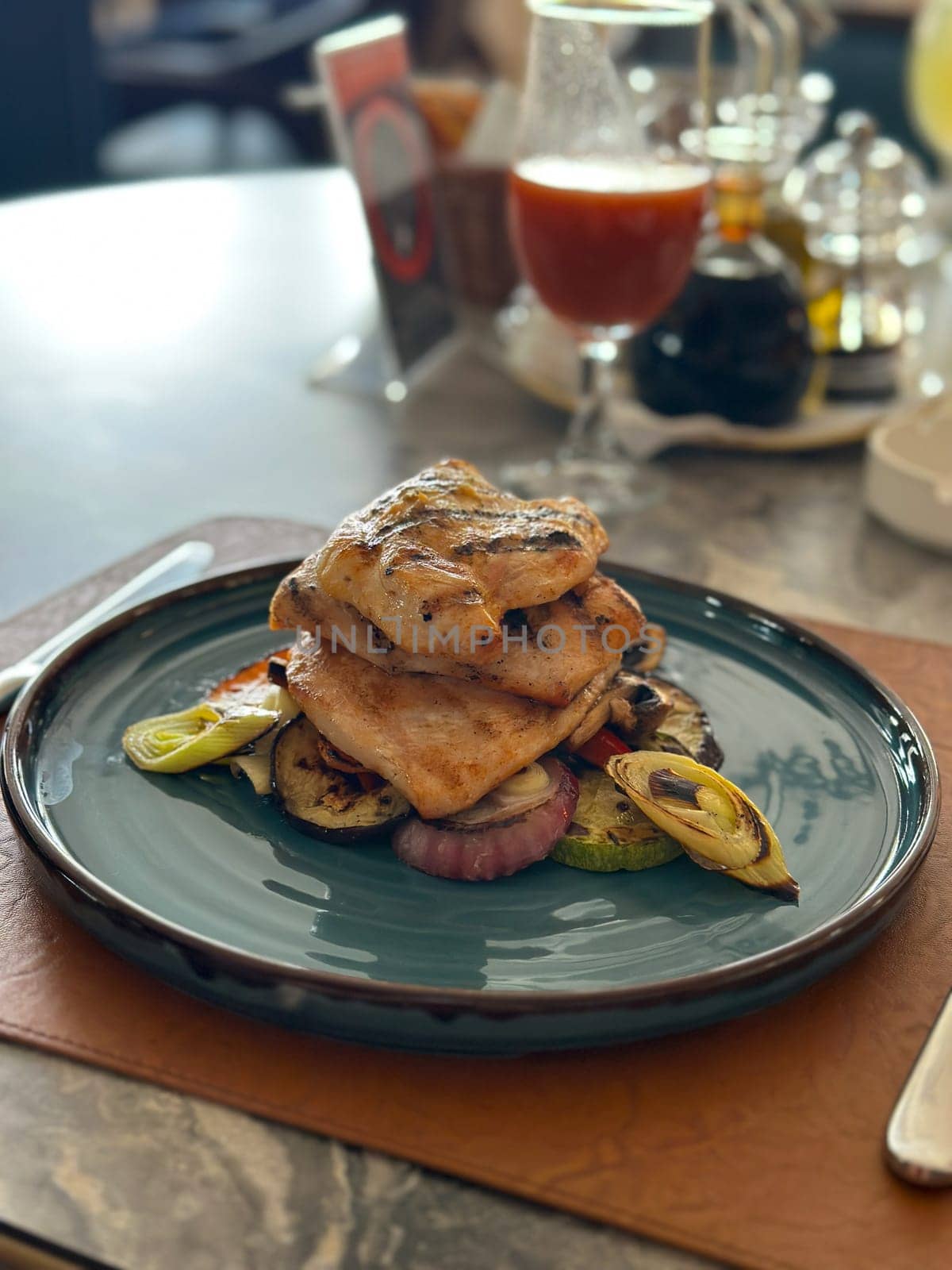 Grilled chicken stacked elegantly on a bed of vegetables, elegantly presented on a blue plate with a glass of juice nearby in restaurant. Culinary scene captures the artistry and appeal of modern dining in a beautifully set table environment