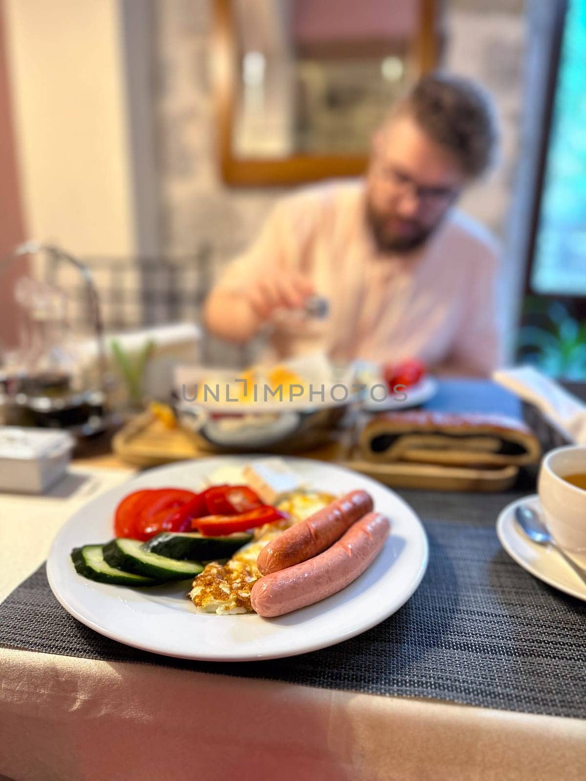 hearty breakfast is beautifully arranged on a plate, featuring sausages, fried eggs, and fresh vegetables. in the background, a person prepares to enjoy this appetizing meal, highlighting a cozy dining.