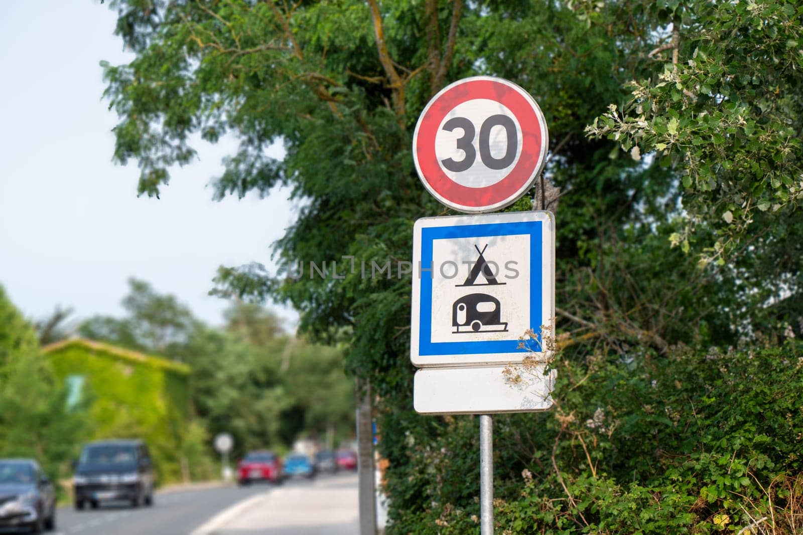 A french sign with a red circle and number 30 . The sign is surrounded by trees and is near a road with cars
