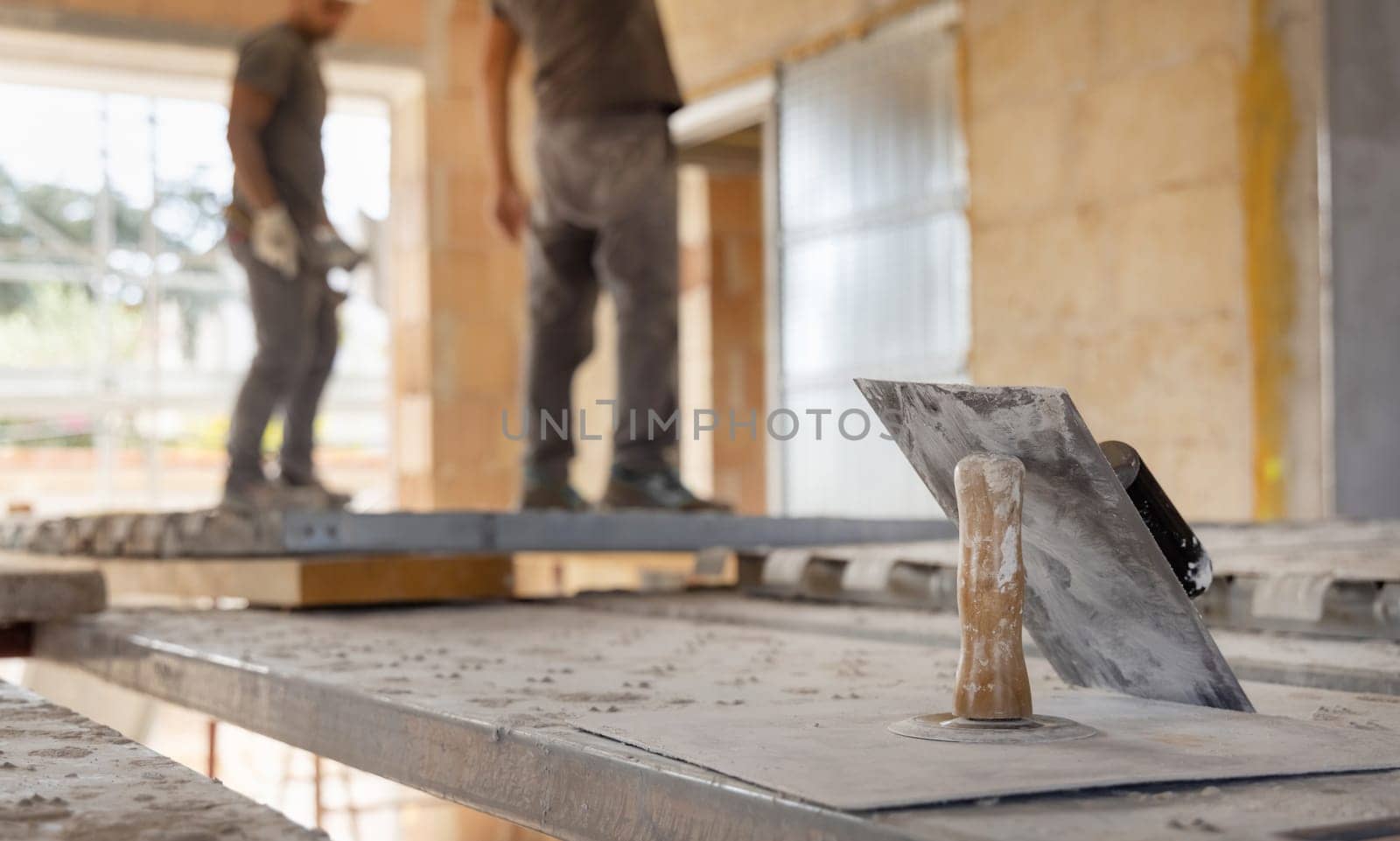 Craftsmen working inside a room standing on scaffoldings, metal trowels in the foreground. Men in grey work clothes during construction or renovation process