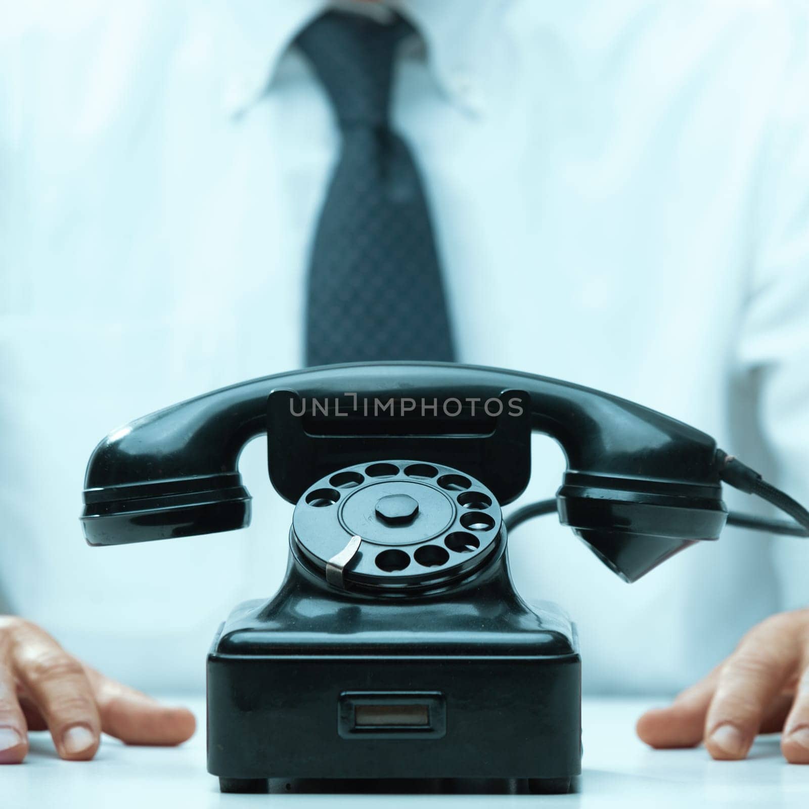 Office worker in a white shirt and tie is sitting at a desk with his hands resting in front of a vintage black telephone