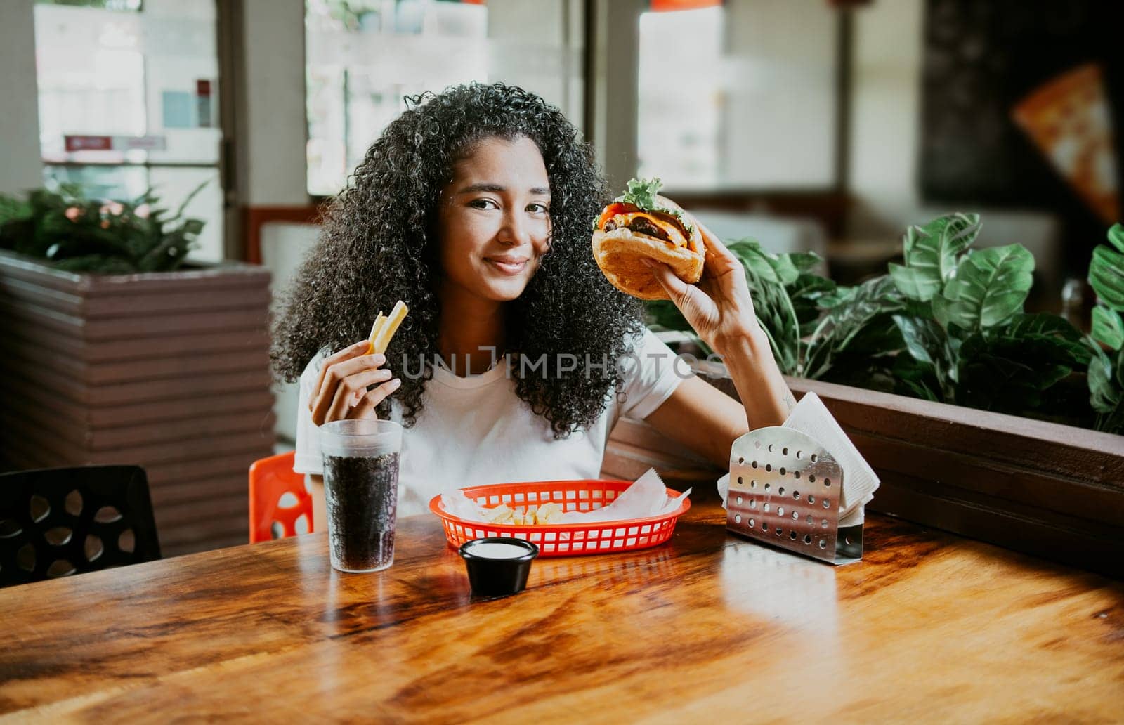 Happy girl enjoying a delicious hamburger sitting in a restaurant. Portrait of young afro woman holding a hamburger with fries in a restaurant