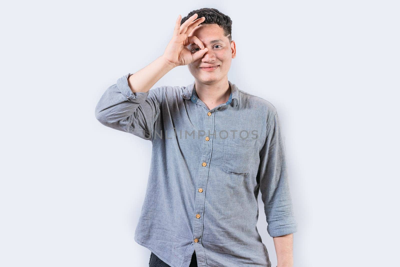 Happy young man making ok gesture with hand looking through fingers. Smiling person making ok gesture looking through fingers isolated