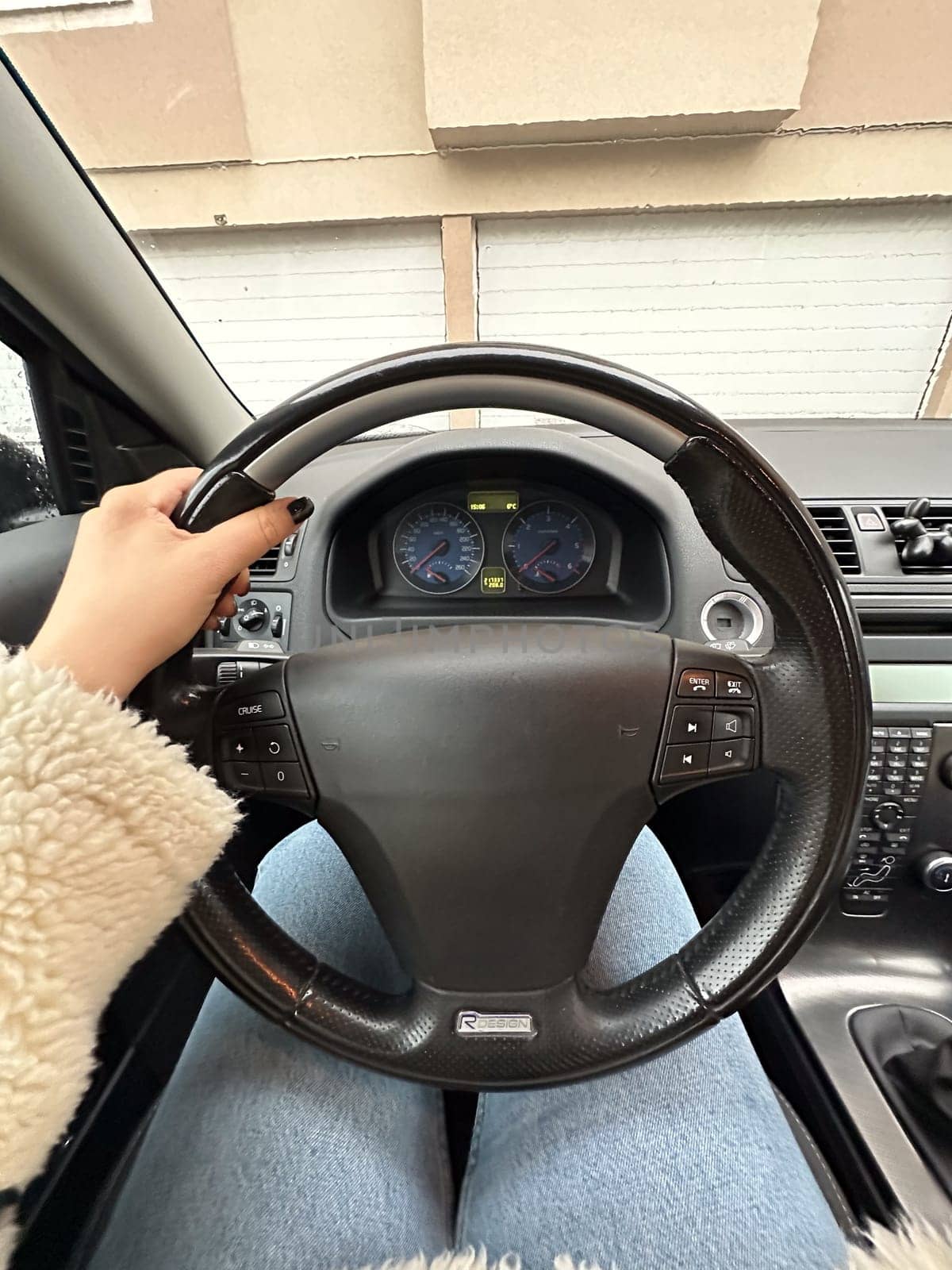 Female hand on a steering wheel inside a modern car, showcasing a casual outfit with a fluffy jacket and jeans. Moment just before starting drive, highlighting dashboard and residential garages outside.