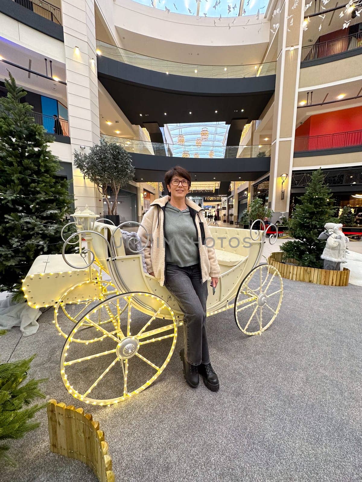 Woman in casual attire standing next to a decorative carriage in a modern shopping mall. Christmas holiday decorations and festive atmosphere create a lively and cheerful scene.
