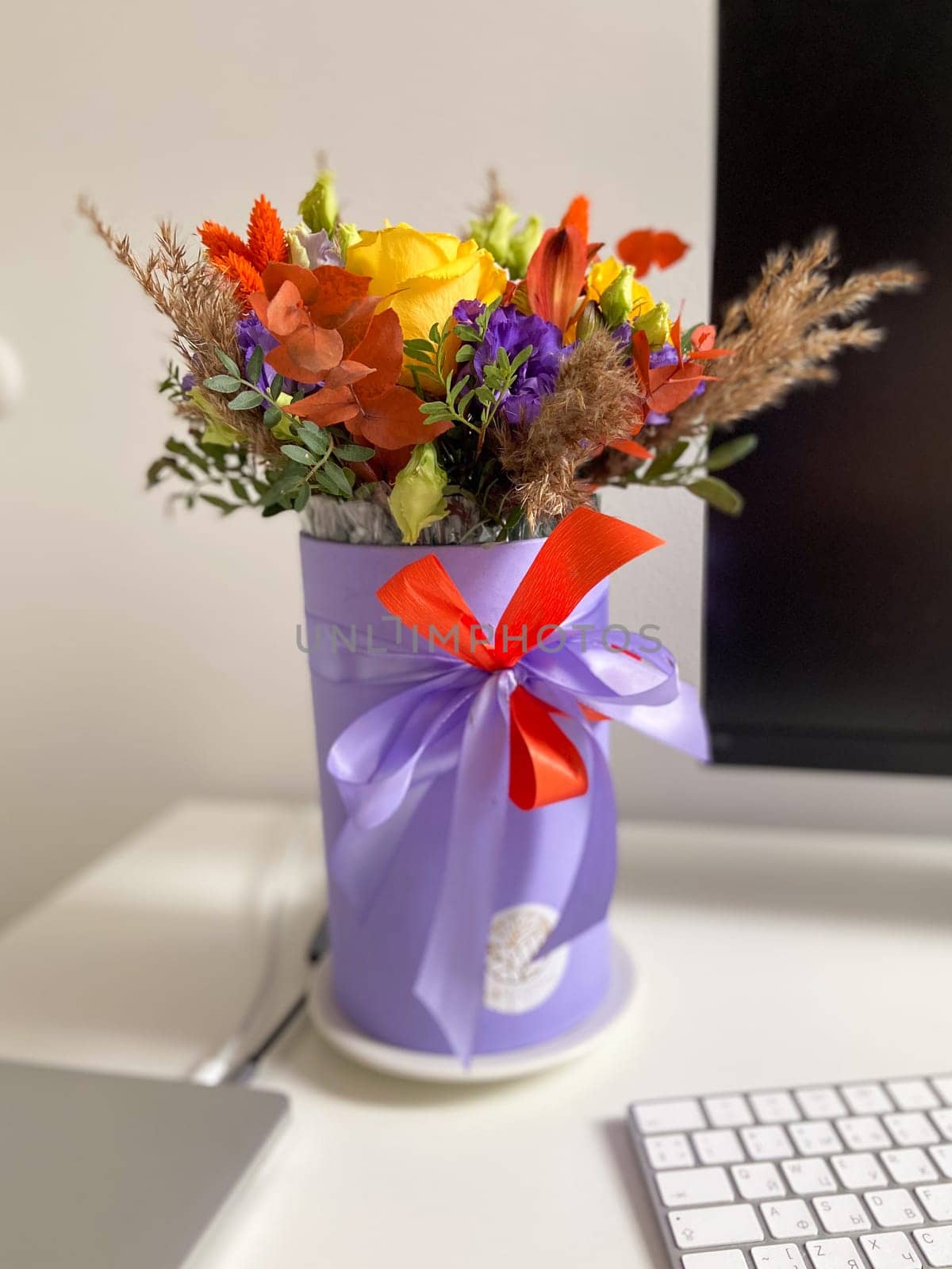Colorful bouquet of flowers in a lavender box standing on a desk, tied with a vibrant red and purple ribbon. Bright, mixed arrangement adds a touch of nature and warmth to modern workspace.