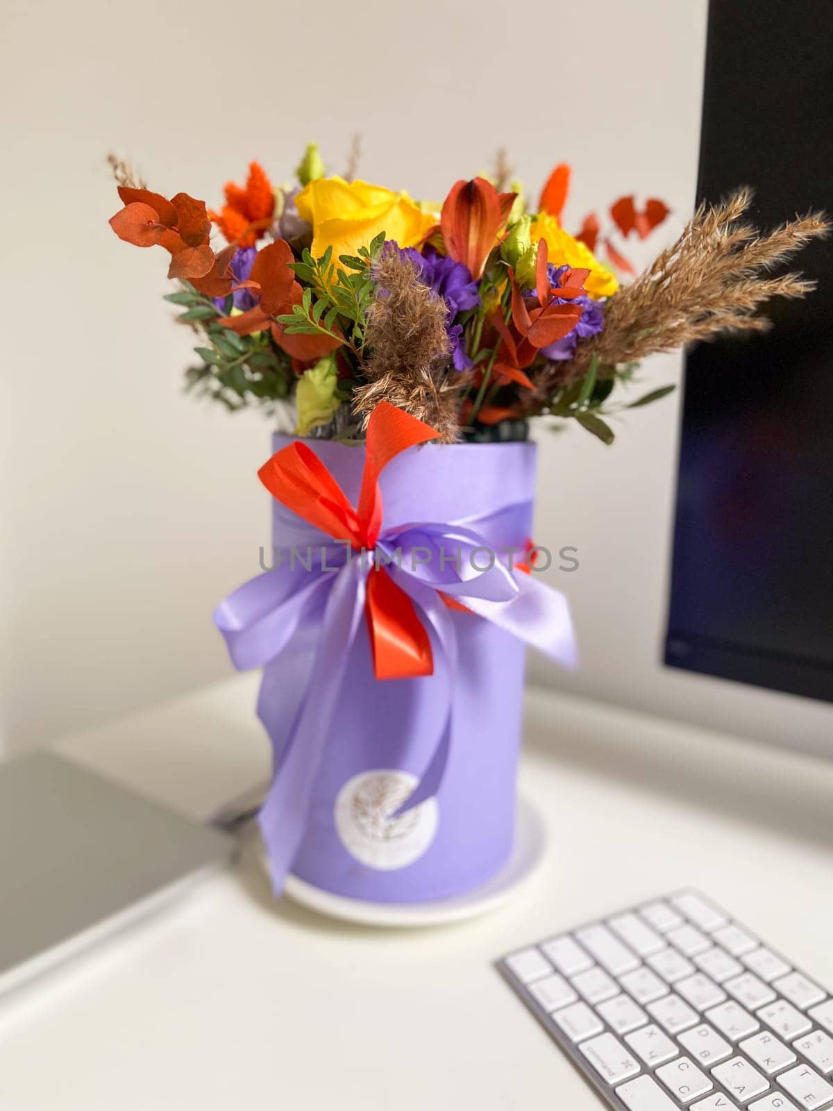 Colorful bouquet of flowers in a lavender box standing on a desk, tied with a vibrant red and purple ribbon. Bright, mixed arrangement adds a touch of nature and warmth to modern workspace.