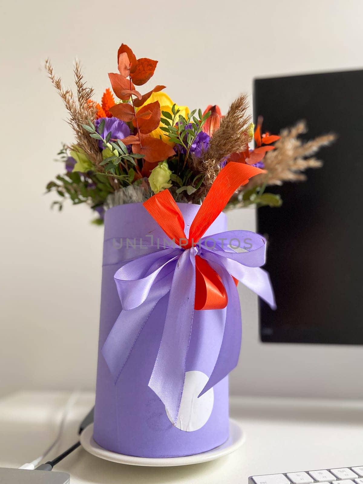 Colorful bouquet of flowers in a lavender box standing on a desk, tied with a vibrant red and purple ribbon. Bright, mixed arrangement adds a touch of nature and warmth to modern workspace.