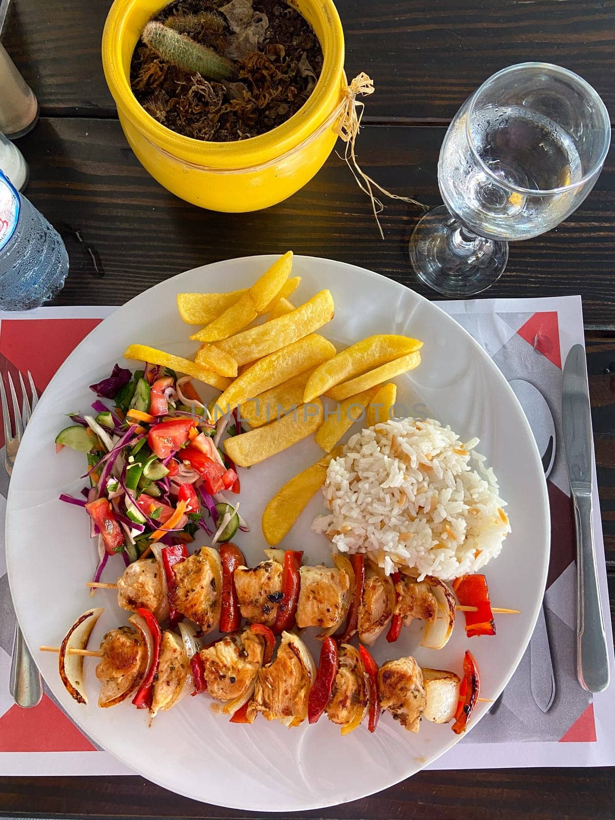 Delicious plate of grilled chicken skewers with rice, salad, and fries on a wooden table setting. Well-balanced and colorful meal, perfect for a vibrant dining experience.