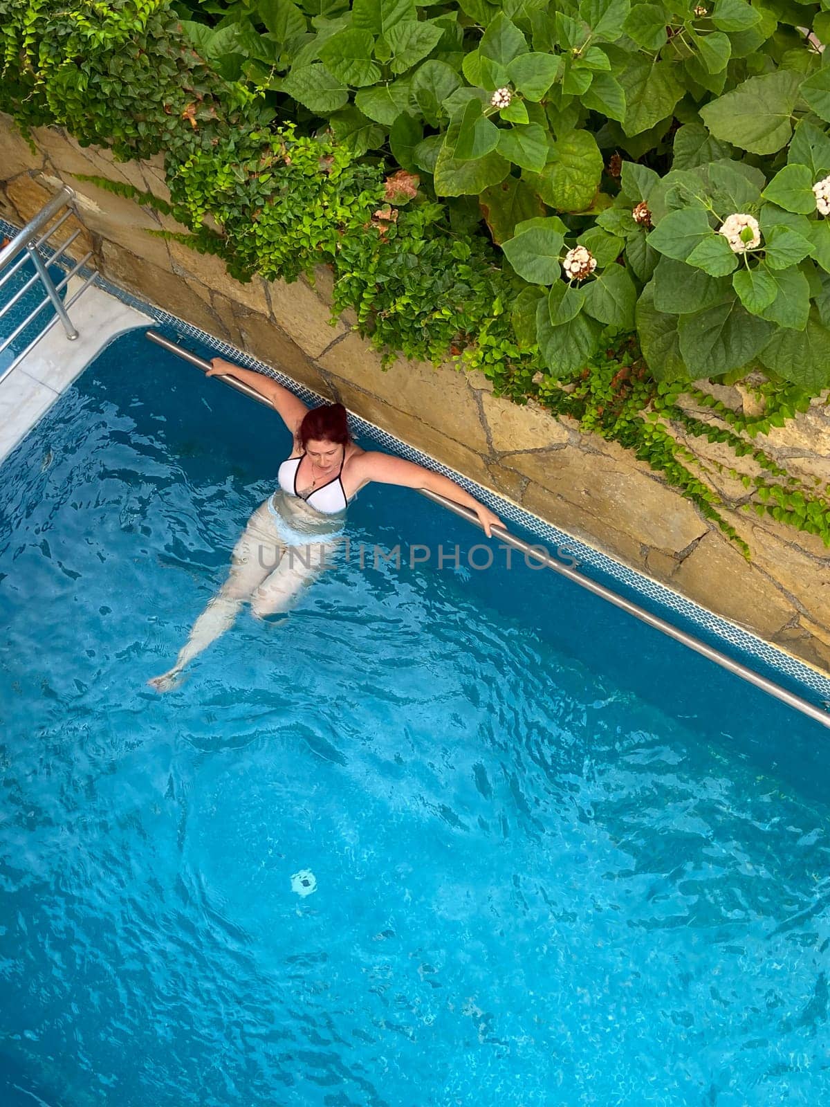 Woman relaxing in a swimming pool near a stone wall adorned with lush green vegetation, enjoying a peaceful moment in the refreshing water. Blend of natural beauty and relaxation in an outdoor setting.