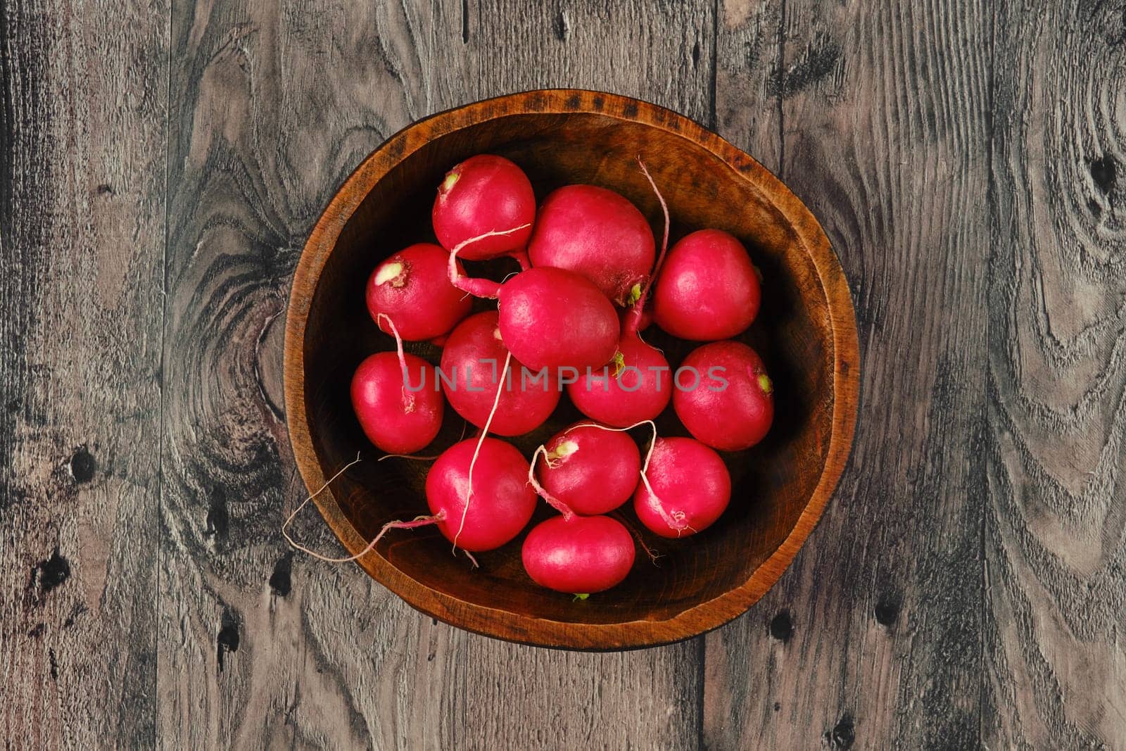 A rustic wooden bowl with vibrant red radishes on a textured, weathered wooden table. The fresh produce showcases the natural beauty of farm-fresh vegetables ready for cooking.