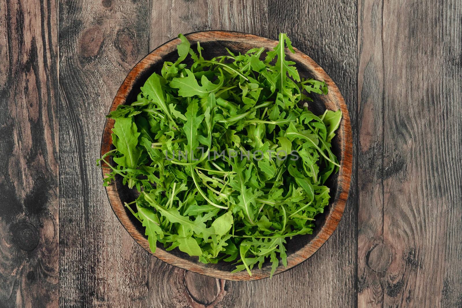 A rustic wooden bowl with fresh arugula leaves on a textured wooden table, showcasing vibrant greenery perfect for salads or garnishes. The natural setting emphasizes freshness.