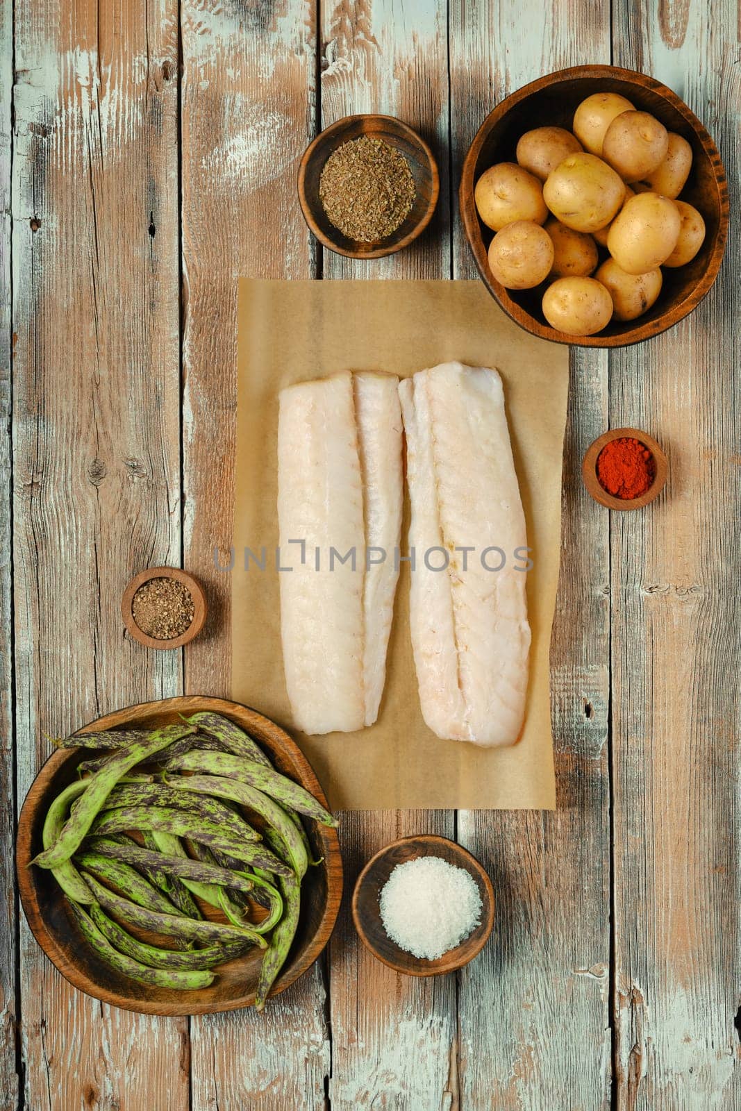Two fresh cod fish fillets on parchment paper surrounded by various spices, green beans, and small potatoes on a wooden table, showcasing a vibrant kitchen arrangement perfect for cooking.
