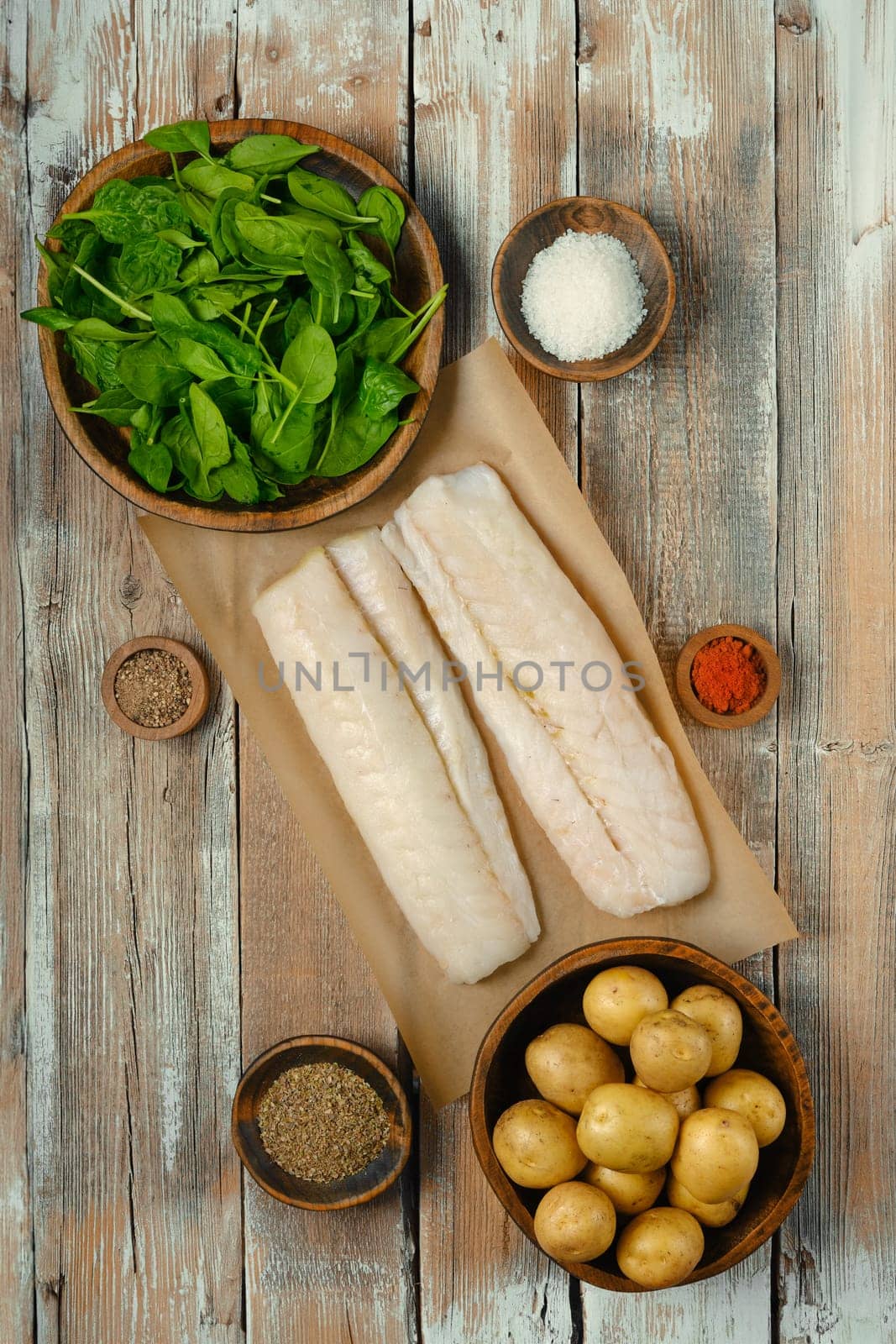 Fresh fish fillets on parchment paper surrounded by bowls of spinach, potatoes, and various spices, ready for meal preparation on a rustic wooden table, perfect for cooking enthusiasts.