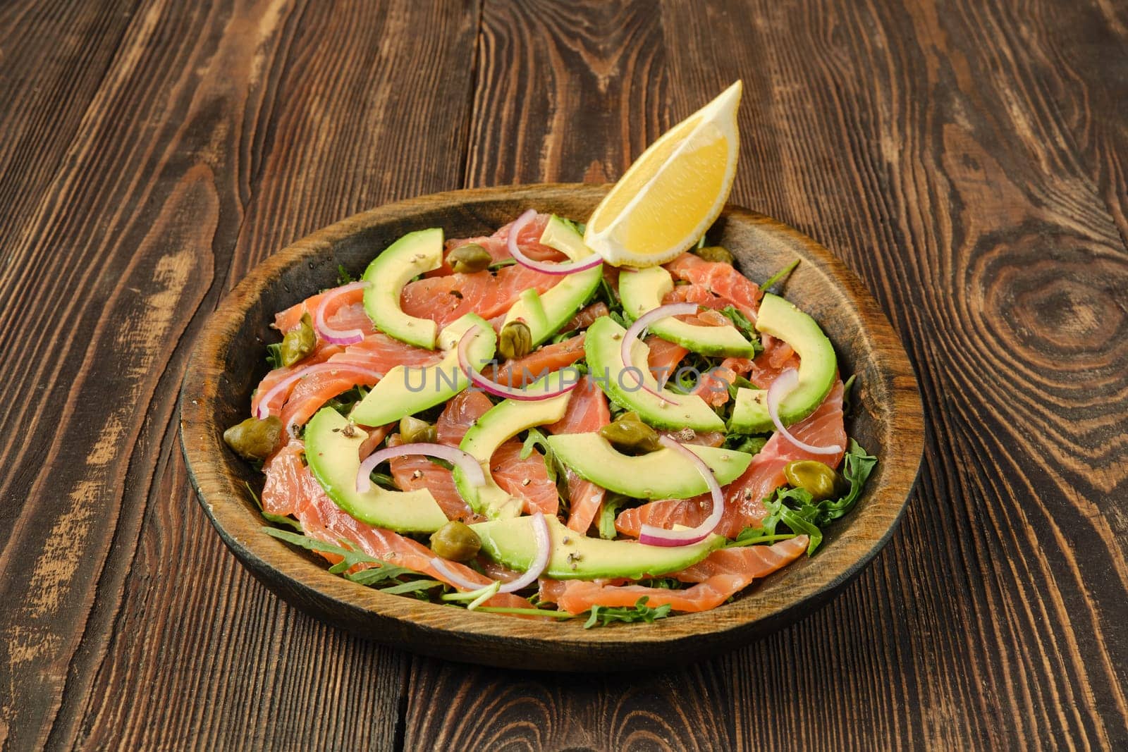 A beautifully arranged plate features slices of salmon topped with avocado, red onion, and capers, garnished with a lemon wedge, set on a rustic wooden table that enhances the dish's appeal.