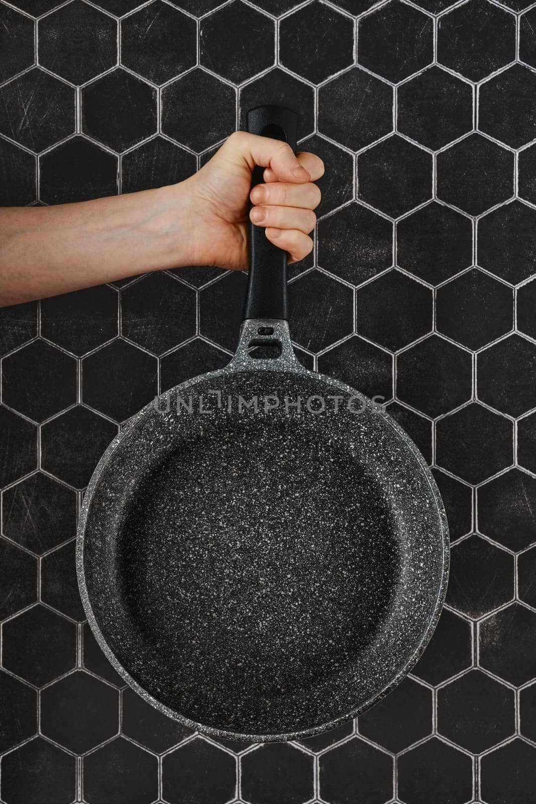 A hand grips a non-stick frying pan with a sturdy handle, positioned against a striking hexagonal dark wall, showcasing kitchen essentials and modern design elements.