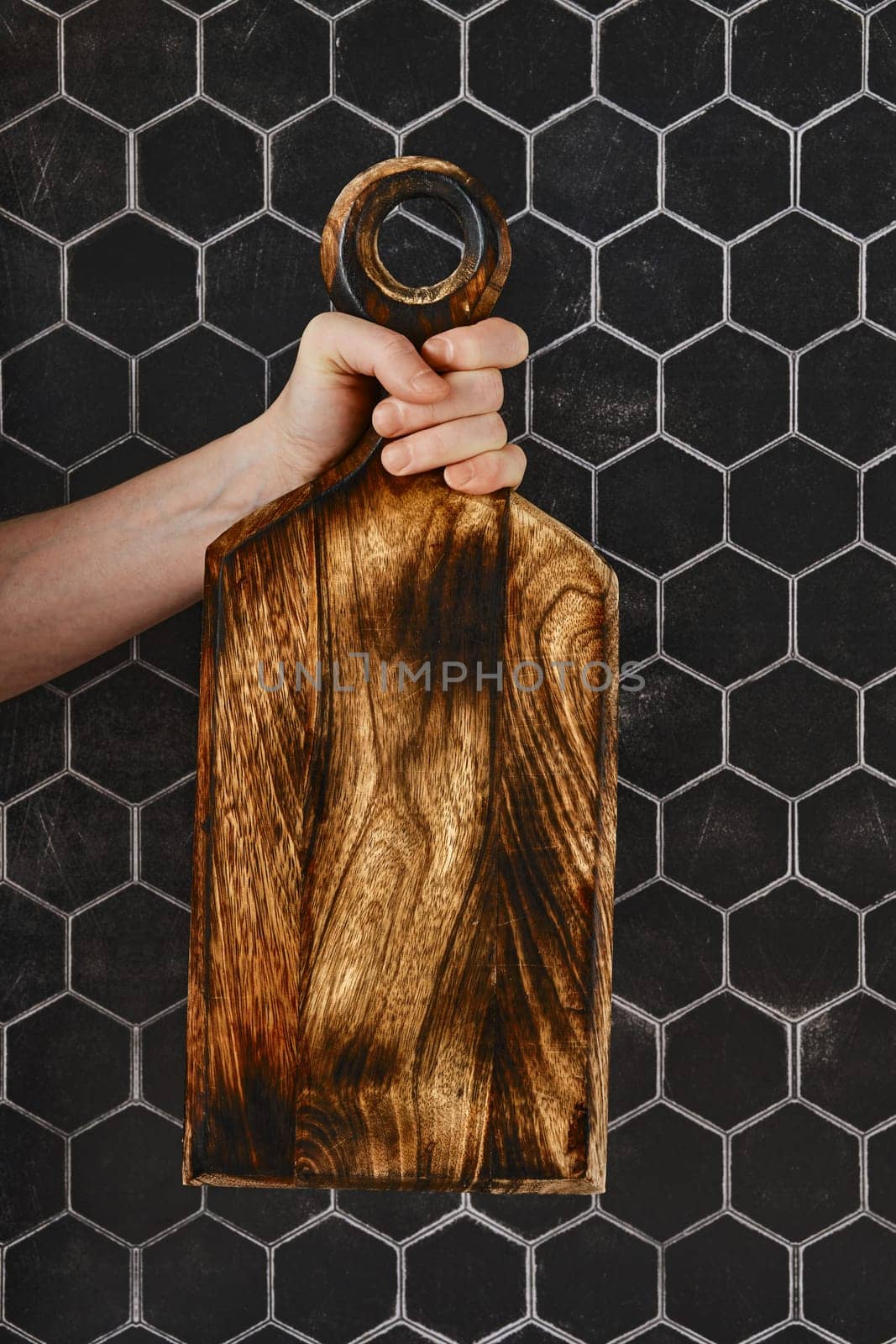 A person grips a stunning wooden cutting board with intricate grain patterns, showcasing craftsmanship. The board is held up against a modern black hexagonal tile wall, adding a stylish touch.
