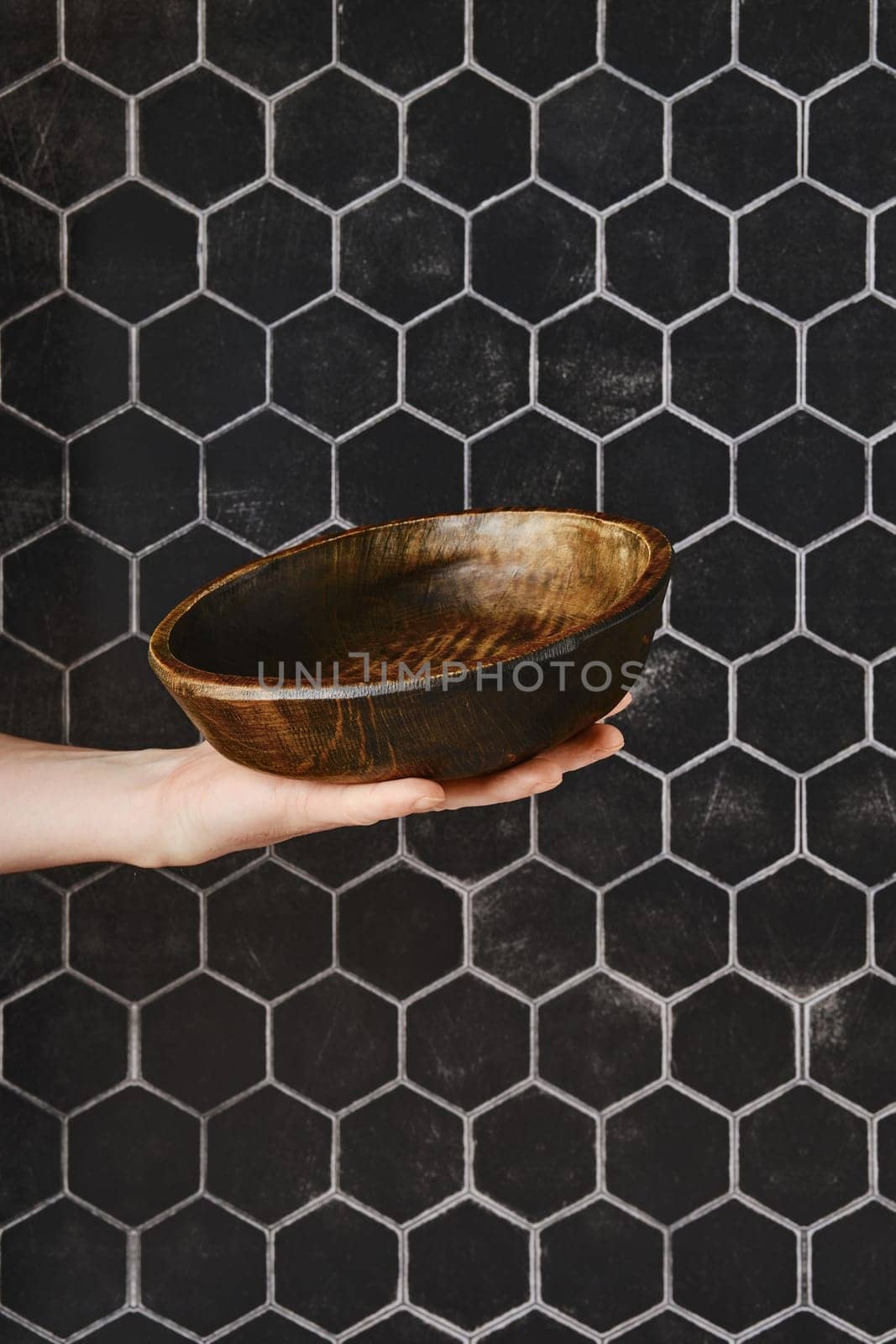 A hand presents a beautifully crafted wooden bowl, showcasing its smooth finish and unique design. The bowl contrasts with a striking hexagonal tile wall, adding elegance to the kitchen setting.