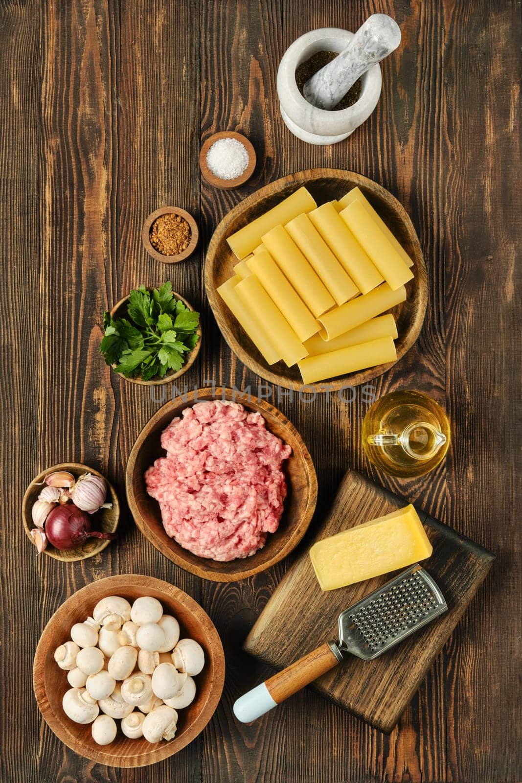 A variety of fresh ingredients is arranged on a wooden surface, featuring ground meat, pasta sheets, mushrooms, cheese, and herbs. The scene showcases preparation for a delicious lasagna