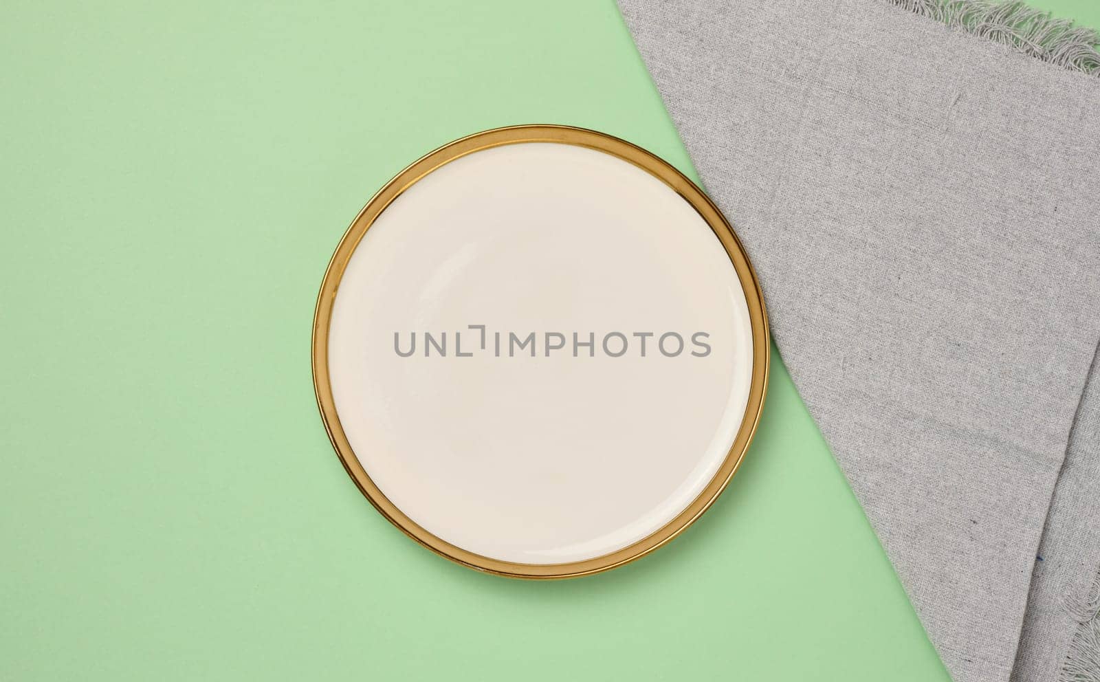 Round ceramic empty white plate and gray linen kitchen towel on green background, top view