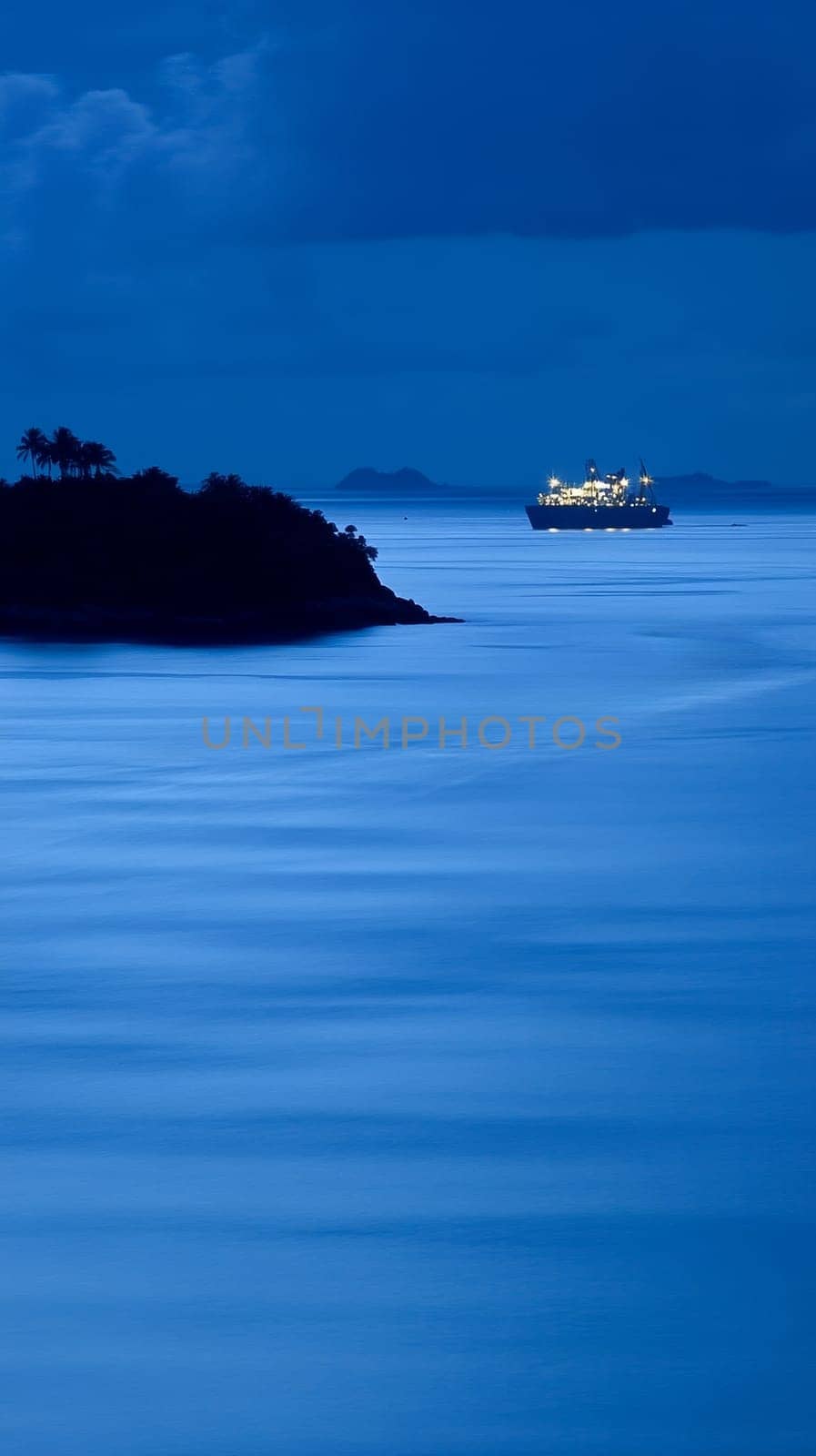 Serene nighttime seascape with an illuminated ship sailing between a silhouetted island and distant mountains, creating a tranquil tropical atmosphere.