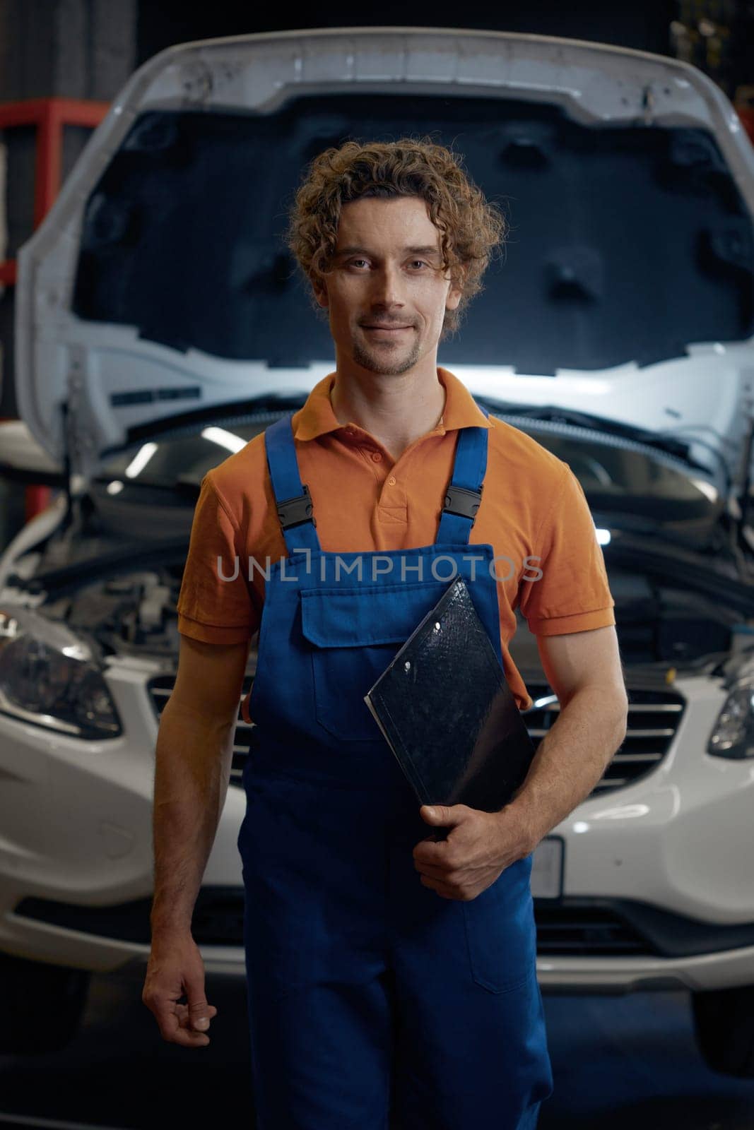 Portrait of young male auto mechanic holding clipboard standing over car with opened hood on repair workshop indoor background