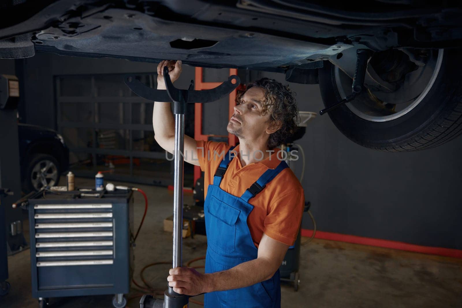 Professional automotive engineer working at car repair station with broken custom vehicle lifted on elevator machine for changing oil