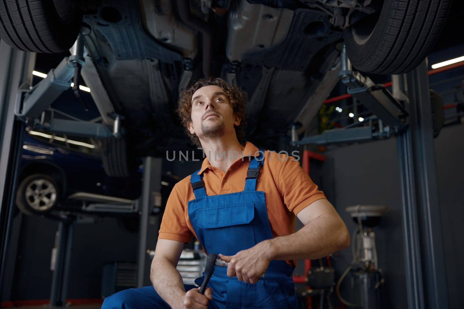 Serious and concentrated man car mechanic holding wrench tool looking at bottom of lifted automobile at repair service station