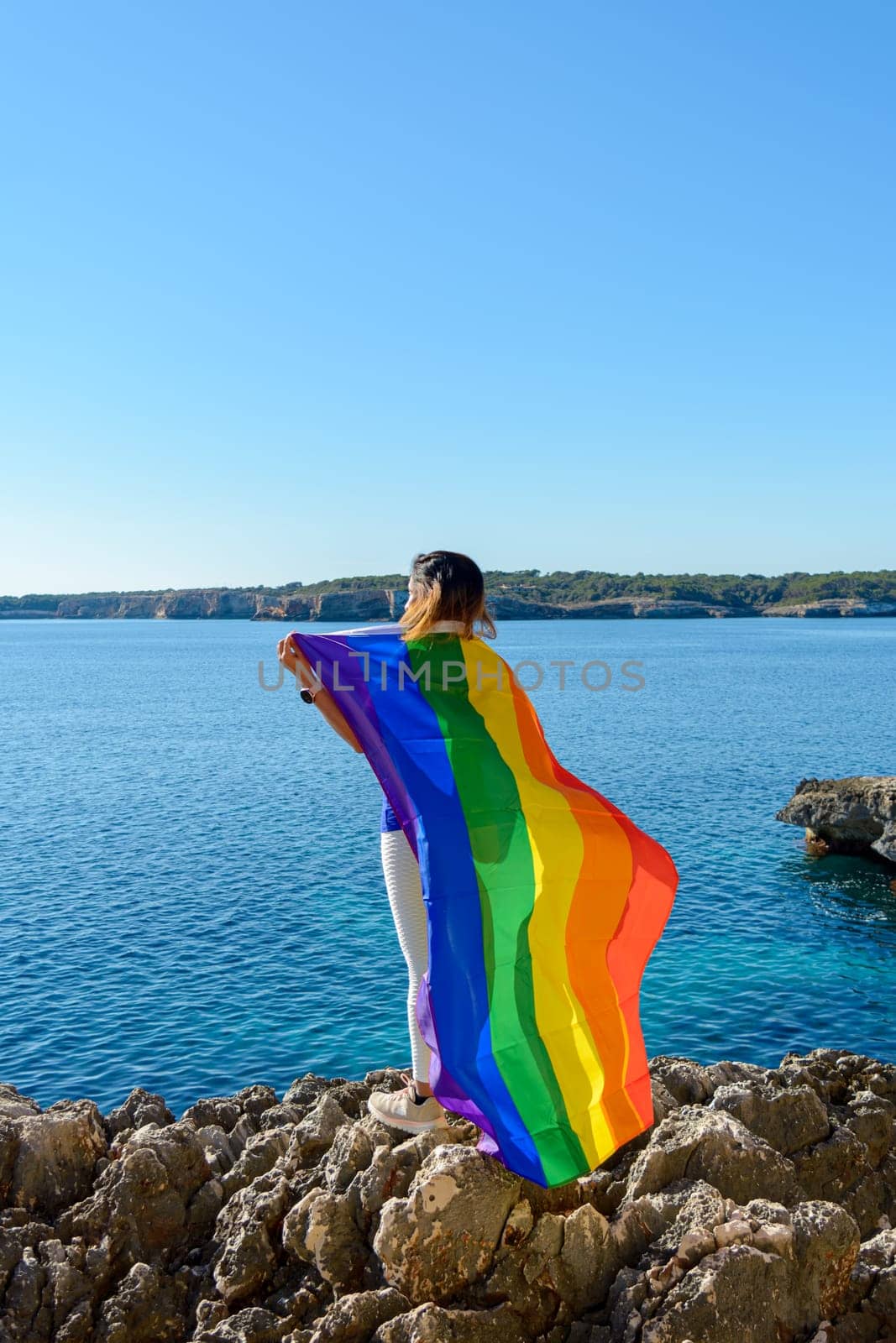 Person on rocky shore holding a colorful rainbow flag against a clear blue sky and ocean backdrop, Woman, middle aged, outdoors waving pride flag, concept of libertat, equality. mallorca, balearic islands, spain