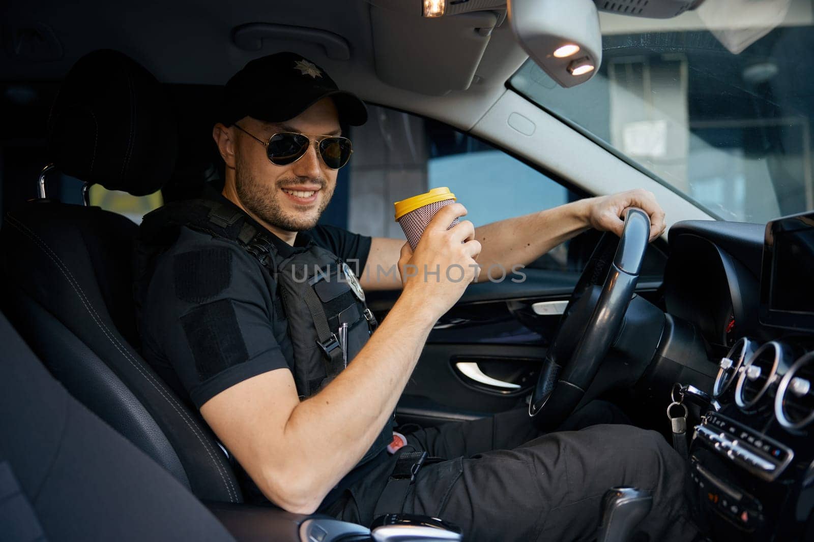 Portrait of smiling patrolman on duty driving car drinking takeaway coffee. Law, order and safe streets concept