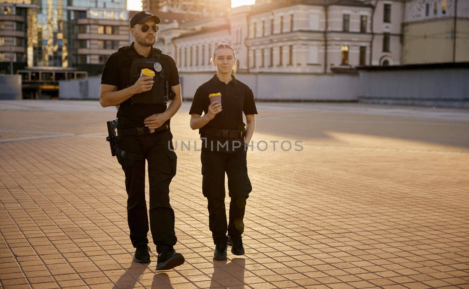 Happy smiling male and female patrol police officers enjoying lunch break. Policeman and policewoman walking down street drinking coffee, eating sandwich and having friendly conversation