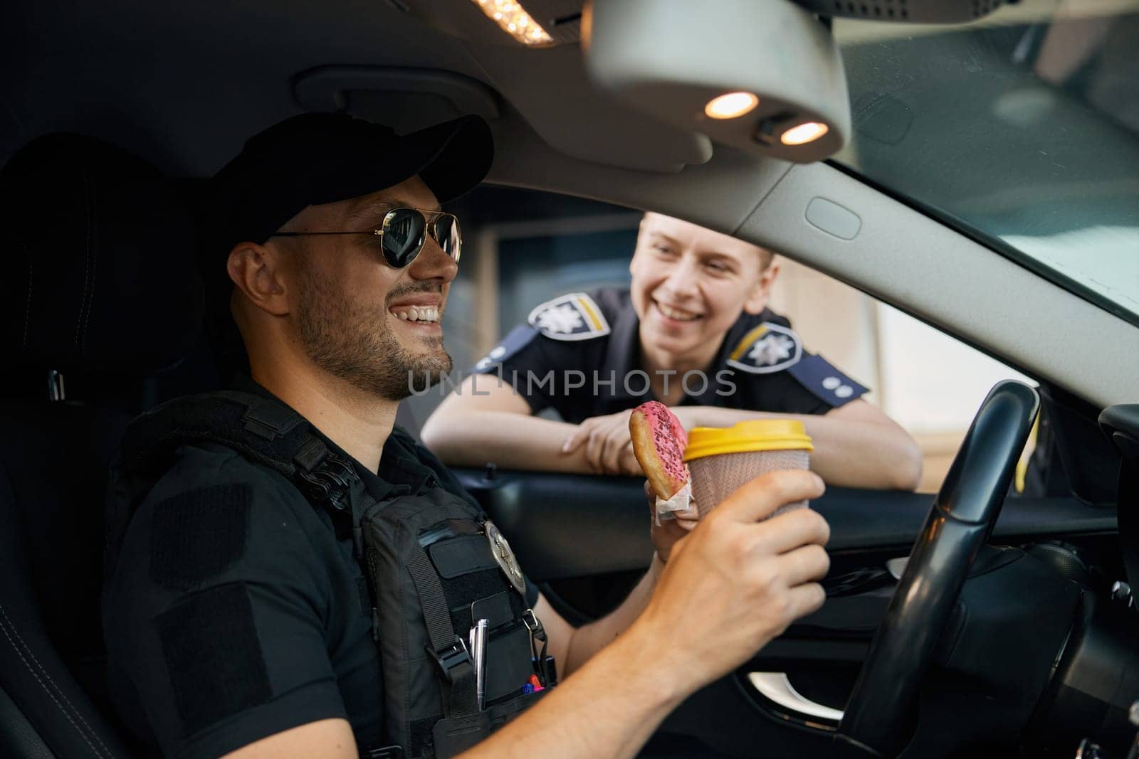 Smiling patrol police officers diverse team sitting in car having coffee break time at work. Policeman with donut and hot aroma drink