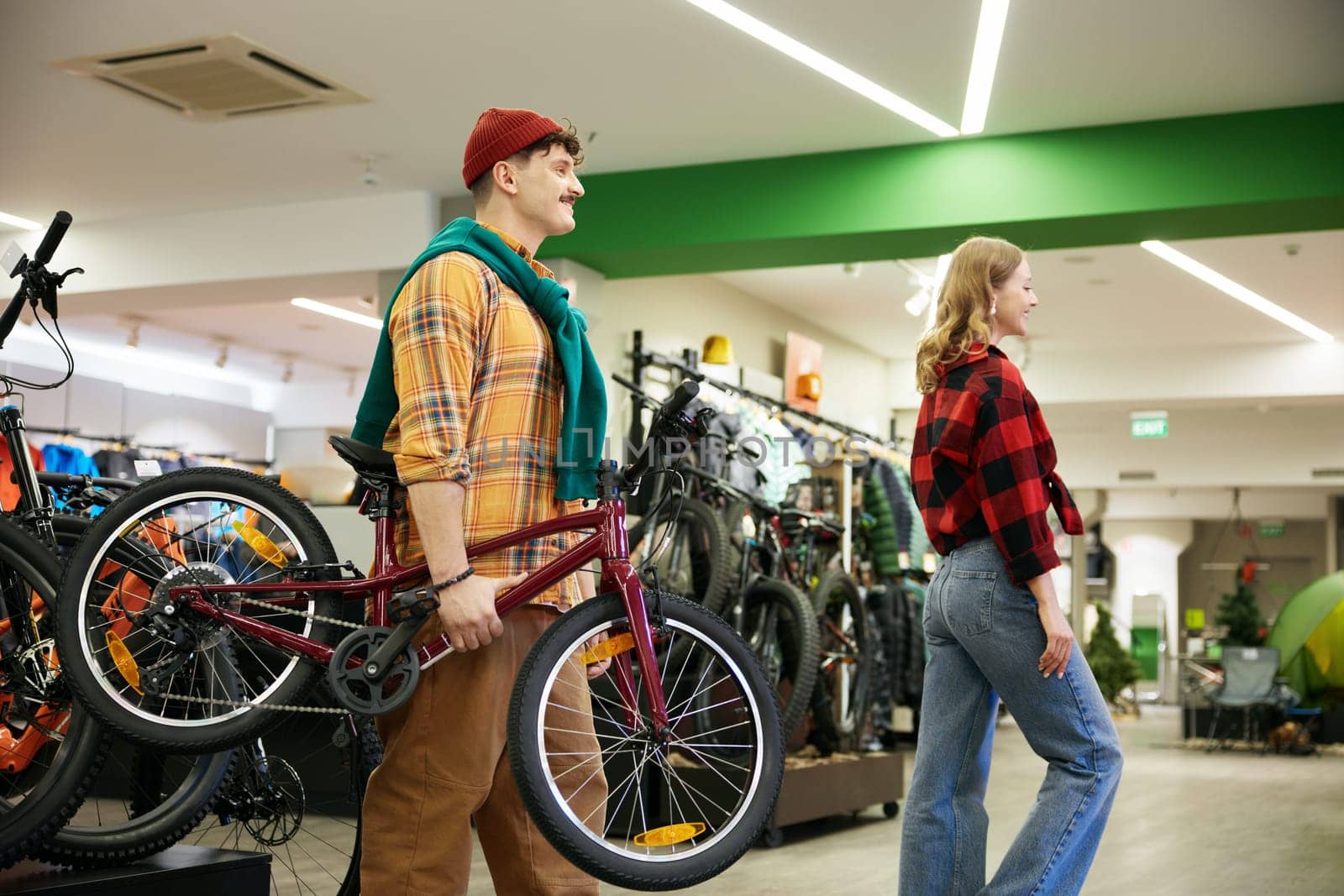 Happy man and woman carrying new kids bike walking through sports shop showcase. Married couple buying new bicycle for their child as birthday gift
