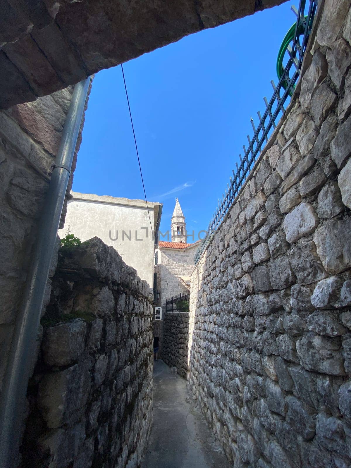 Narrow stone alleyway leading to a historic tower against a clear blue sky, capturing charm and mystery of ancient architecture. Confined passage and towering structures evoke a sense of exploration and timeless beauty.