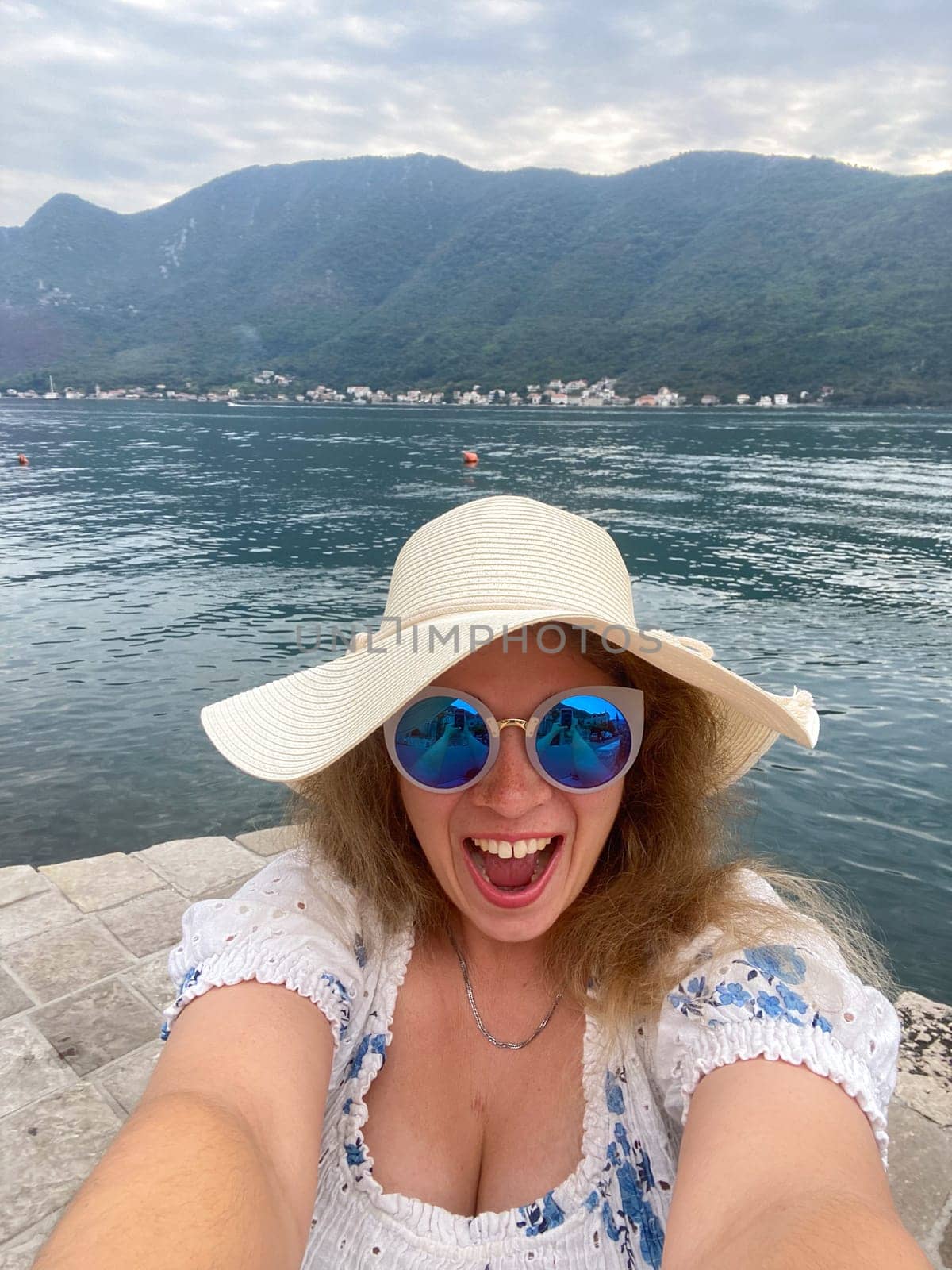 Funny woman in a sun hat and sunglasses smiles joyfully by a serene lake with majestic mountains in background. Taking selfie with moment of happiness and natural beauty during her vacation.