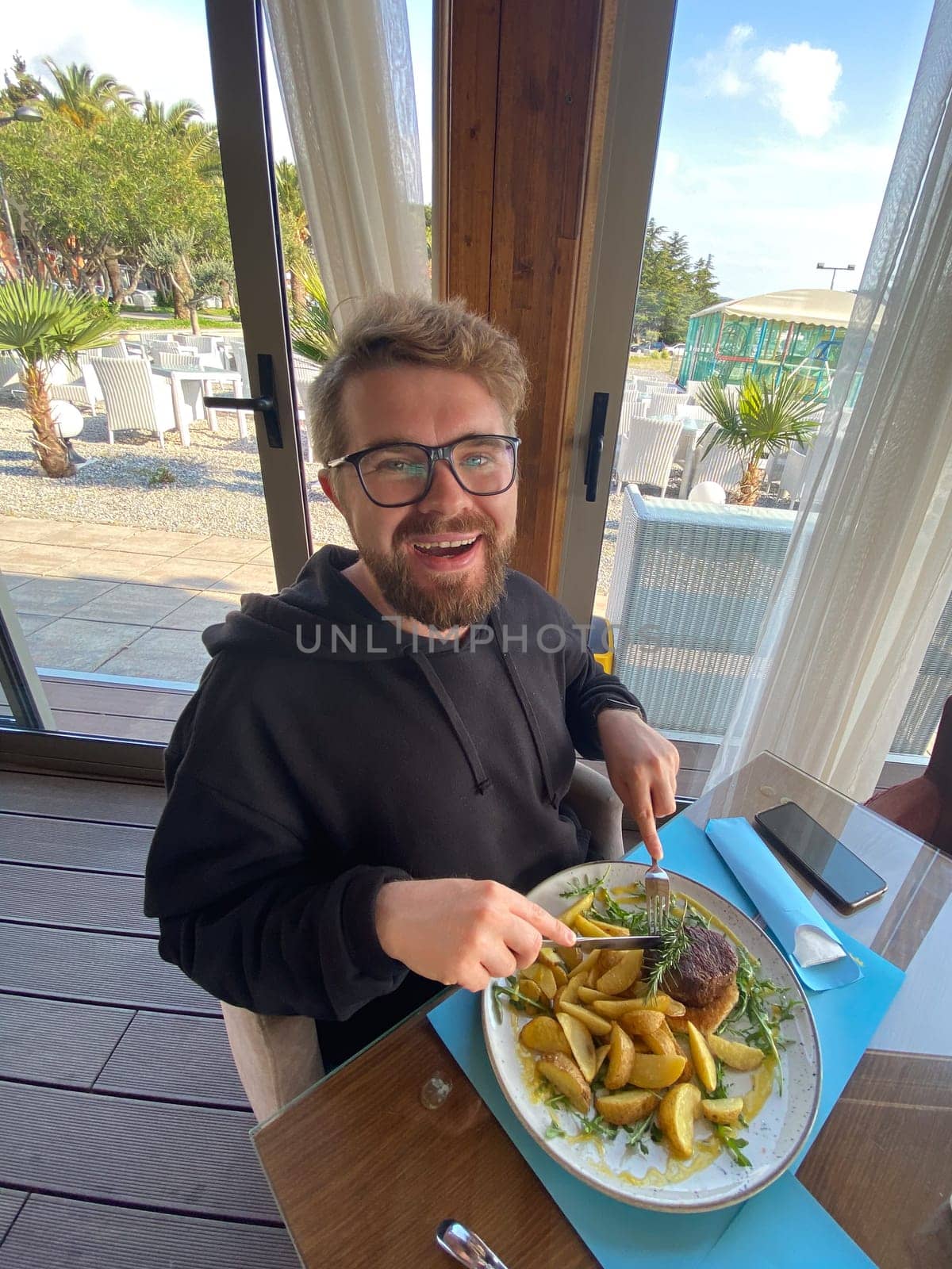Bearded man enjoys a meal at a seaside restaurant, with a plate of steak and potatoes. Bright and open atmosphere, surrounded by palm trees and a scenic view of the beach outside.