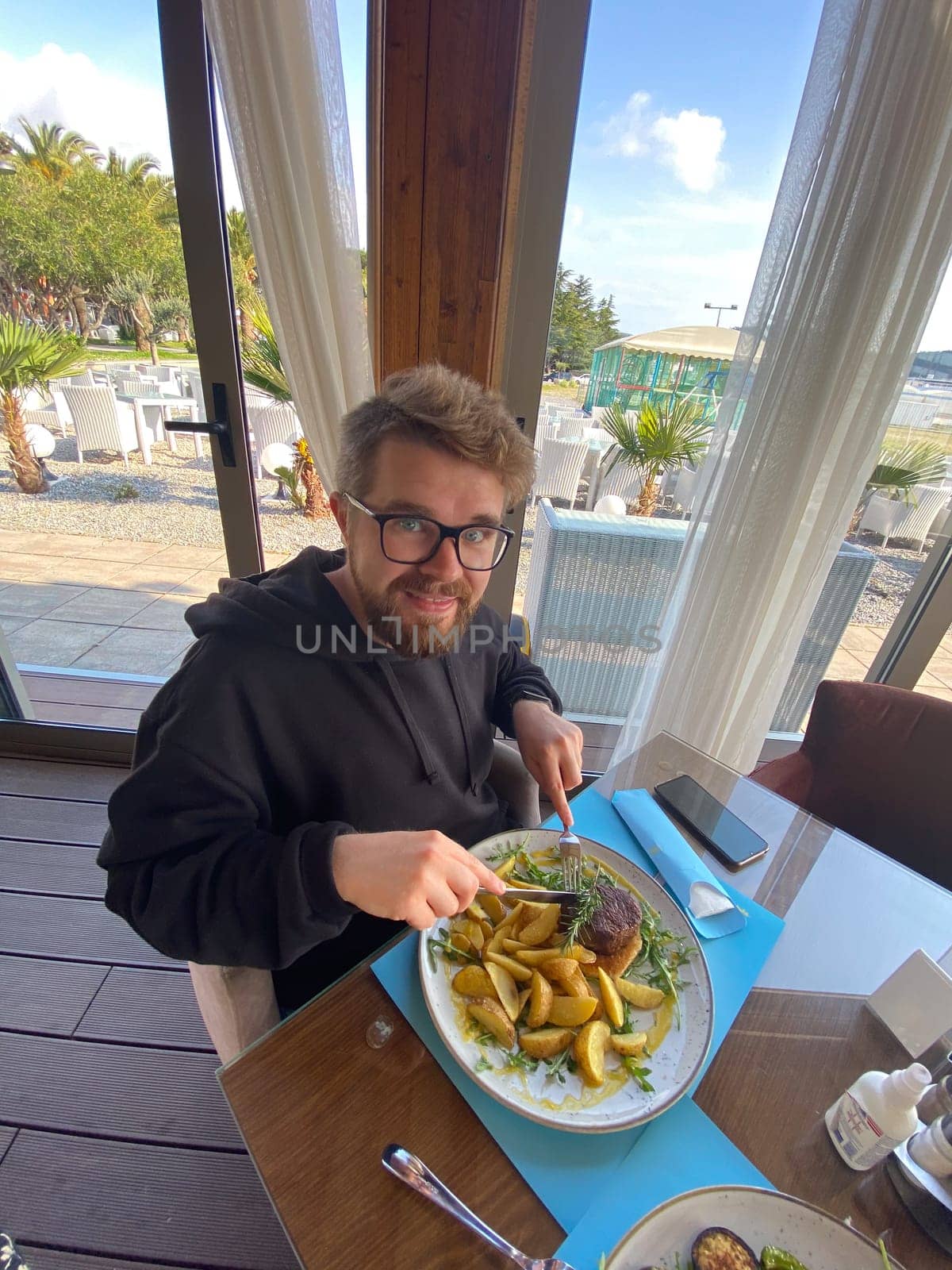 Bearded man enjoys a meal at a seaside restaurant, with a plate of steak and potatoes. Bright and open atmosphere, surrounded by palm trees and a scenic view of the beach outside.