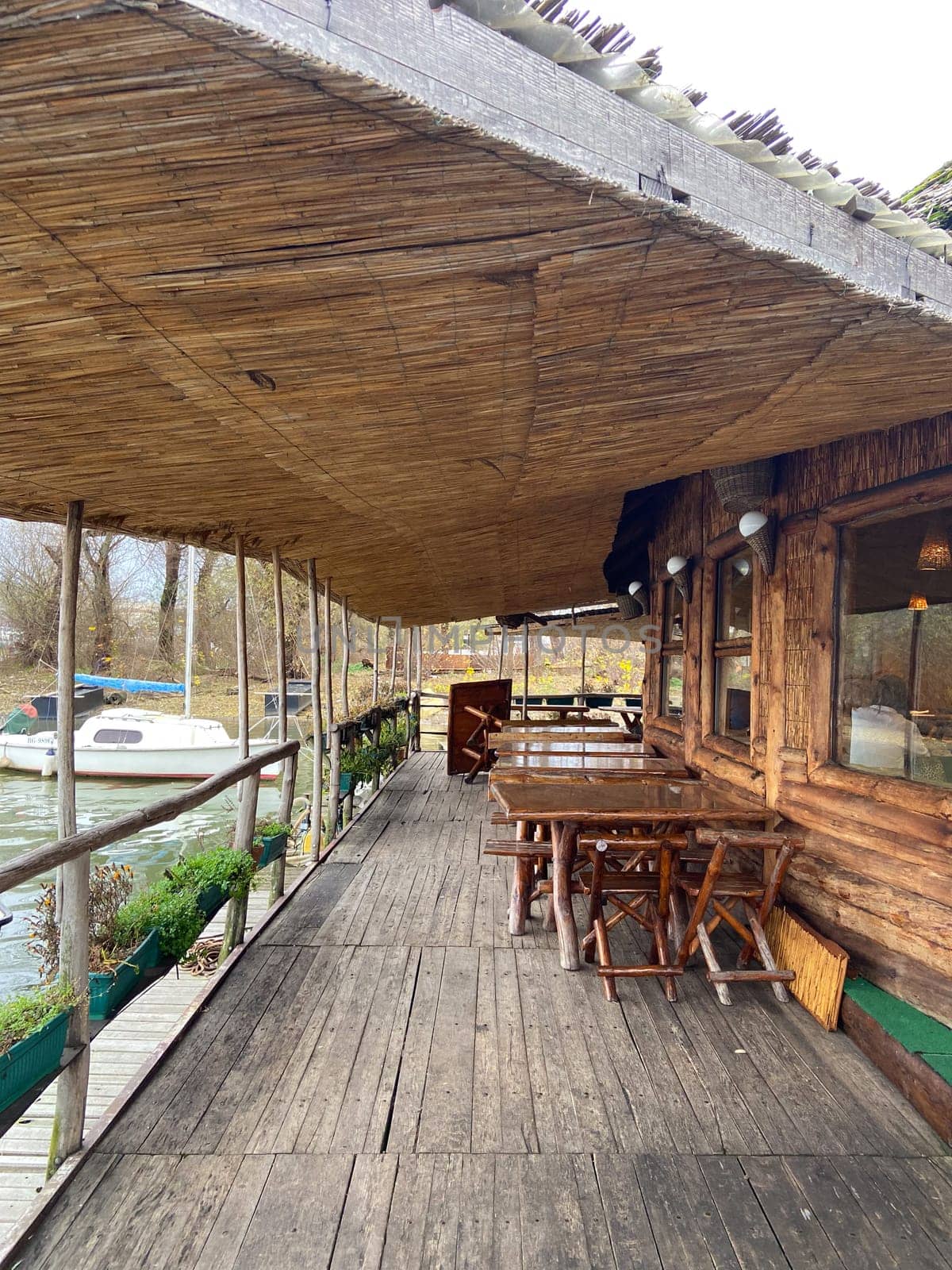 Rustic waterfront deck with wooden tables and a thatched roof overlooks serene river. Outdoor seating area offers a tranquil place to relax and enjoy the natural surroundings.
