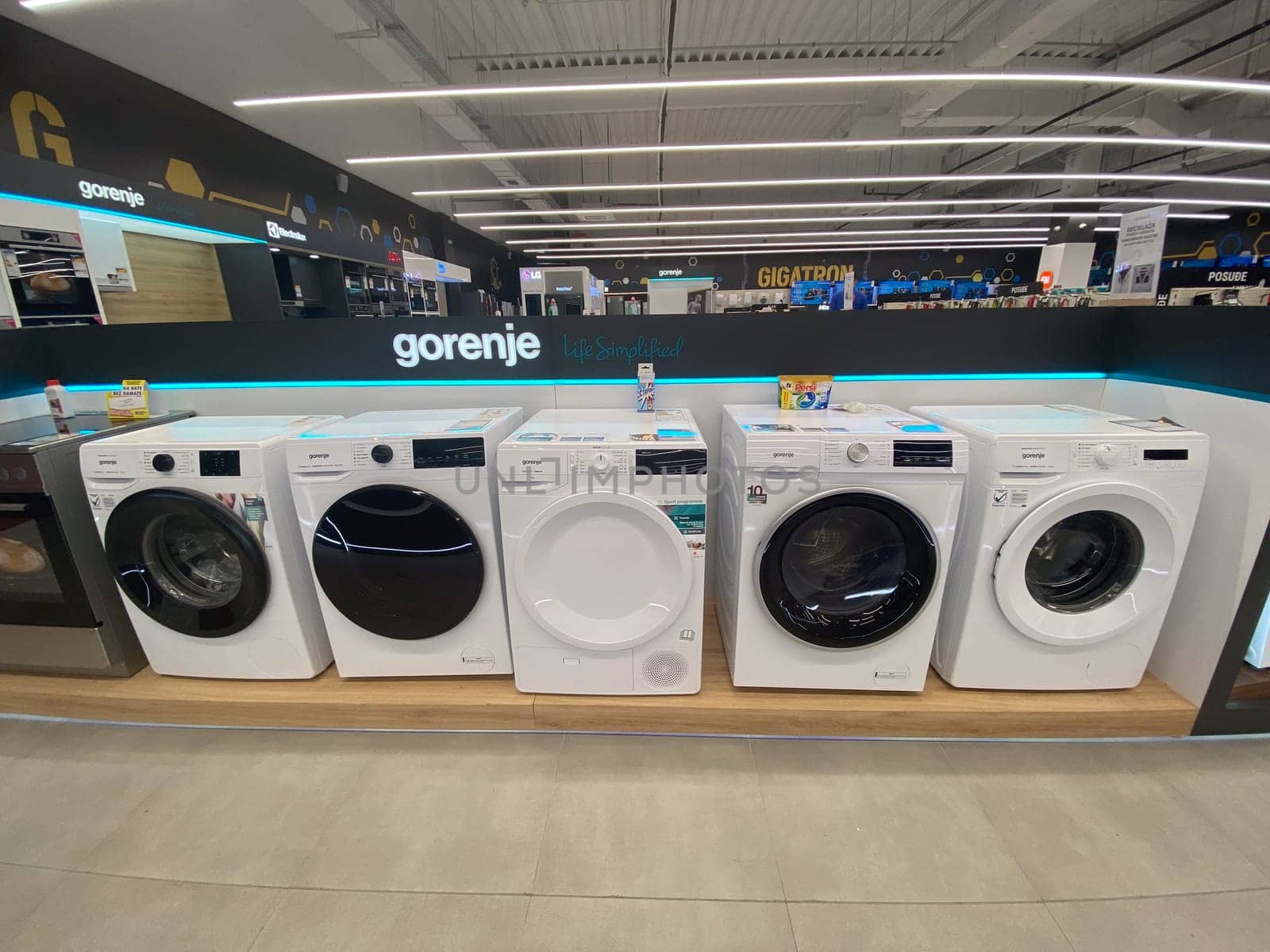 Row of modern washing machines is displayed in an electronics store, showcasing the latest models with sleek designs and advanced features. Under bright lights, the machines stand out against the store contemporary interior.