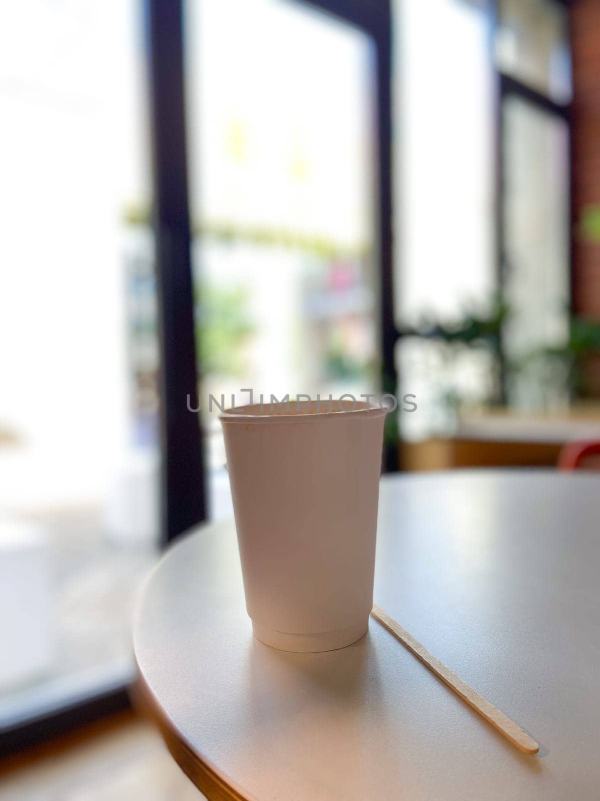 Simple white paper cup sits on a round table in a cozy cafe, inviting a moment of relaxation and warmth. Blurred background suggests a peaceful indoor setting, perfect for enjoying a quiet coffee break