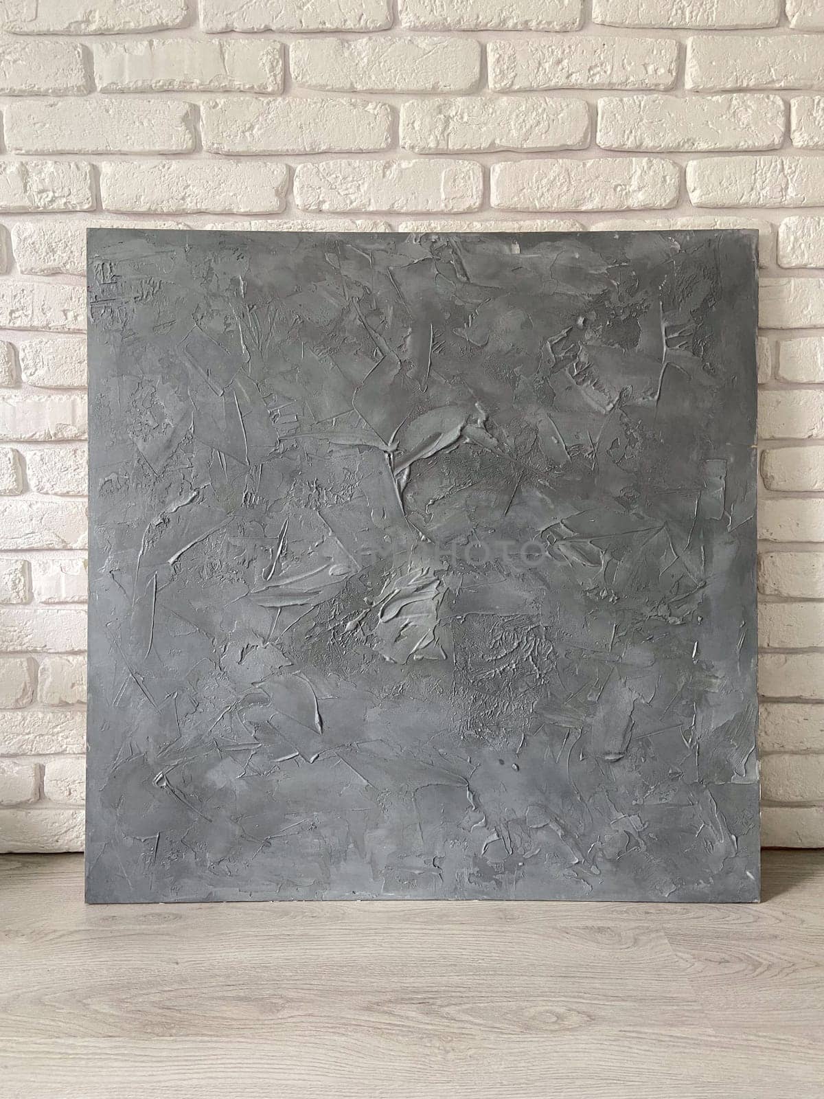 Textured gray canvas leans against a white brick wall, offering a stark contrast and highlighting the intricate details within the artwork. Artistic blend of the canvas meets the structured simplicity of the urban backdrop.