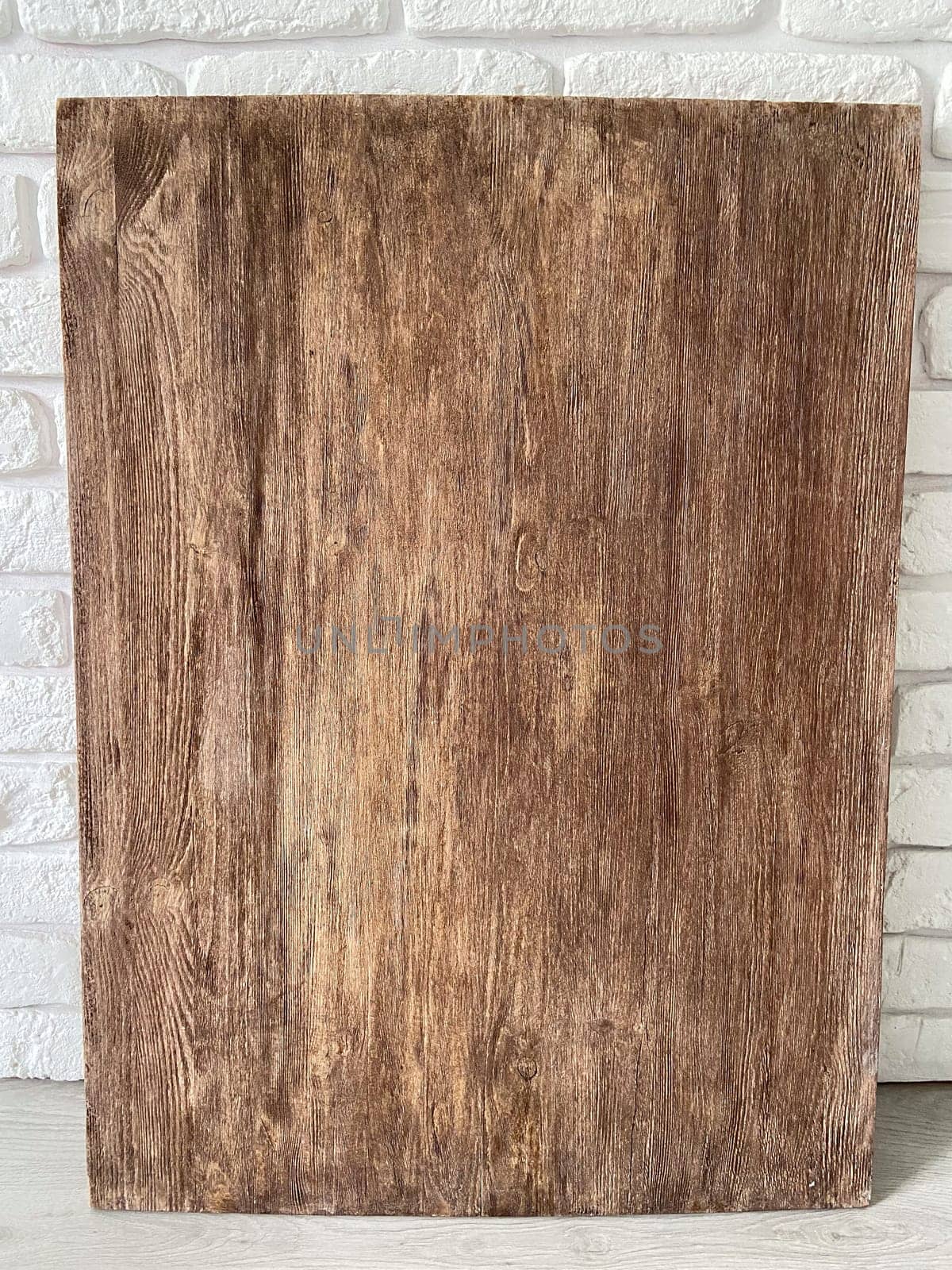 Rustic wooden panel leans gracefully against a textured white brick wall, showcasing its rich grain patterns and natural warmth. Contrast between the organic wood and the structured brick provides a harmonious blend of nature and architecture.