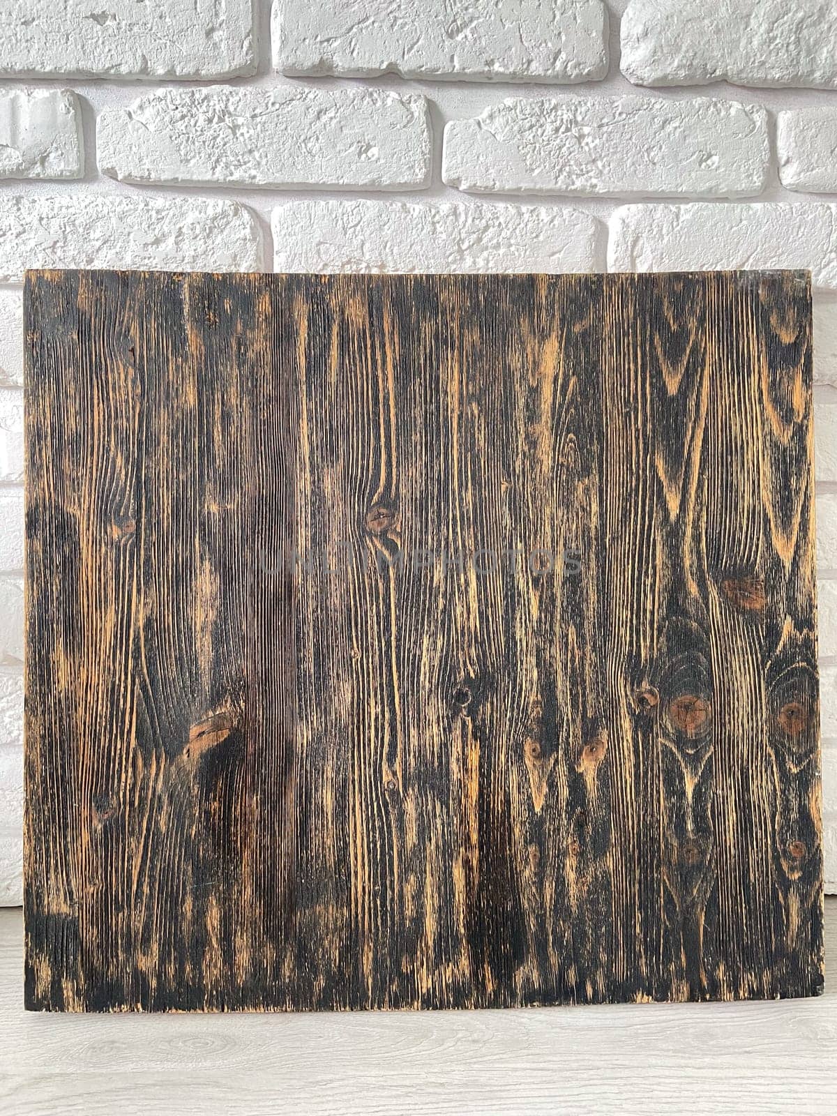 Rustic wooden panel leans gracefully against a textured white brick wall, showcasing its rich grain patterns and natural warmth. Contrast between the organic wood and the structured brick provides a harmonious blend of nature and architecture.