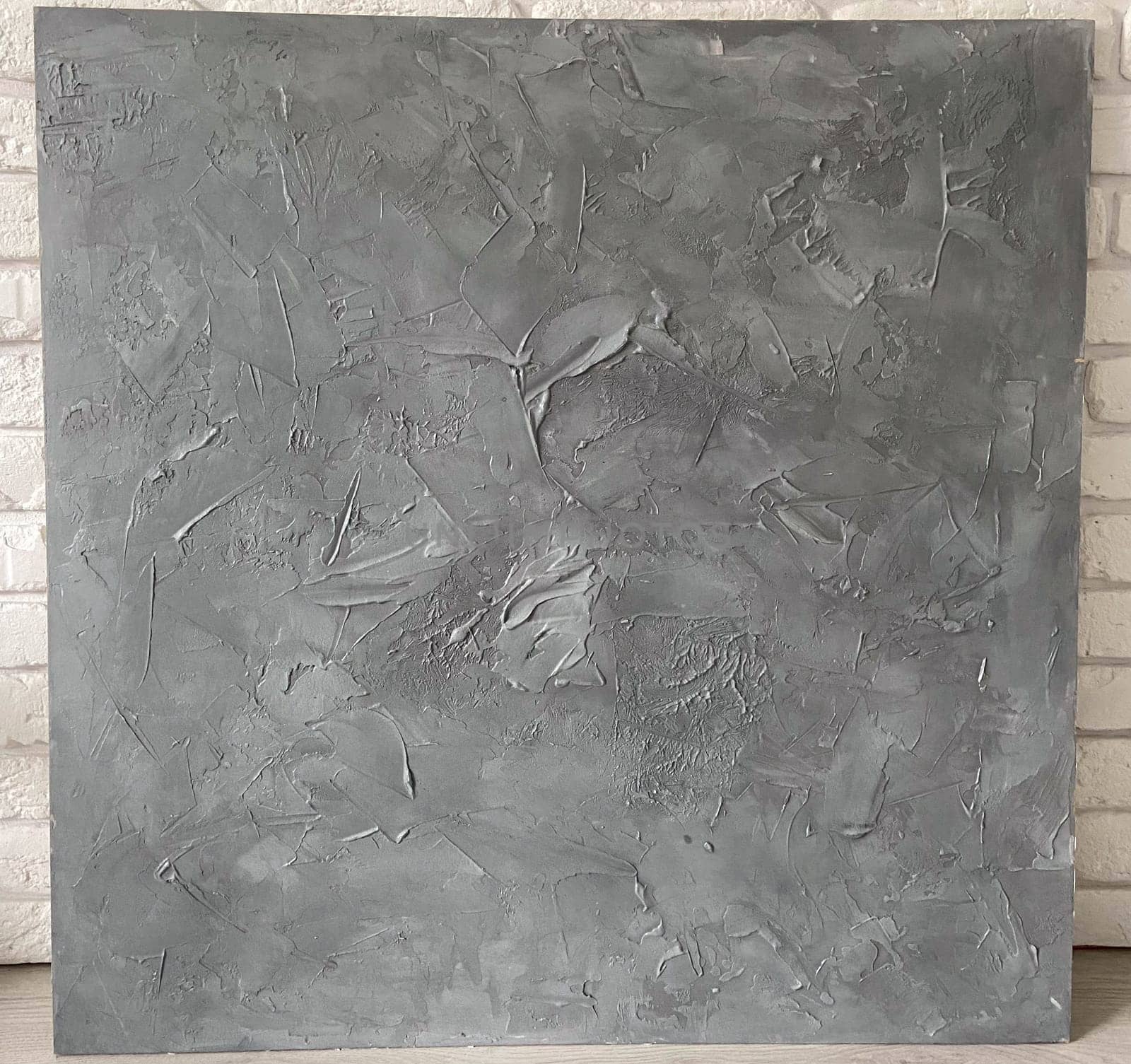Textured gray canvas leans against a white brick wall, offering a stark contrast and highlighting the intricate details within the artwork. Artistic blend of the canvas meets the structured simplicity of the urban backdrop.