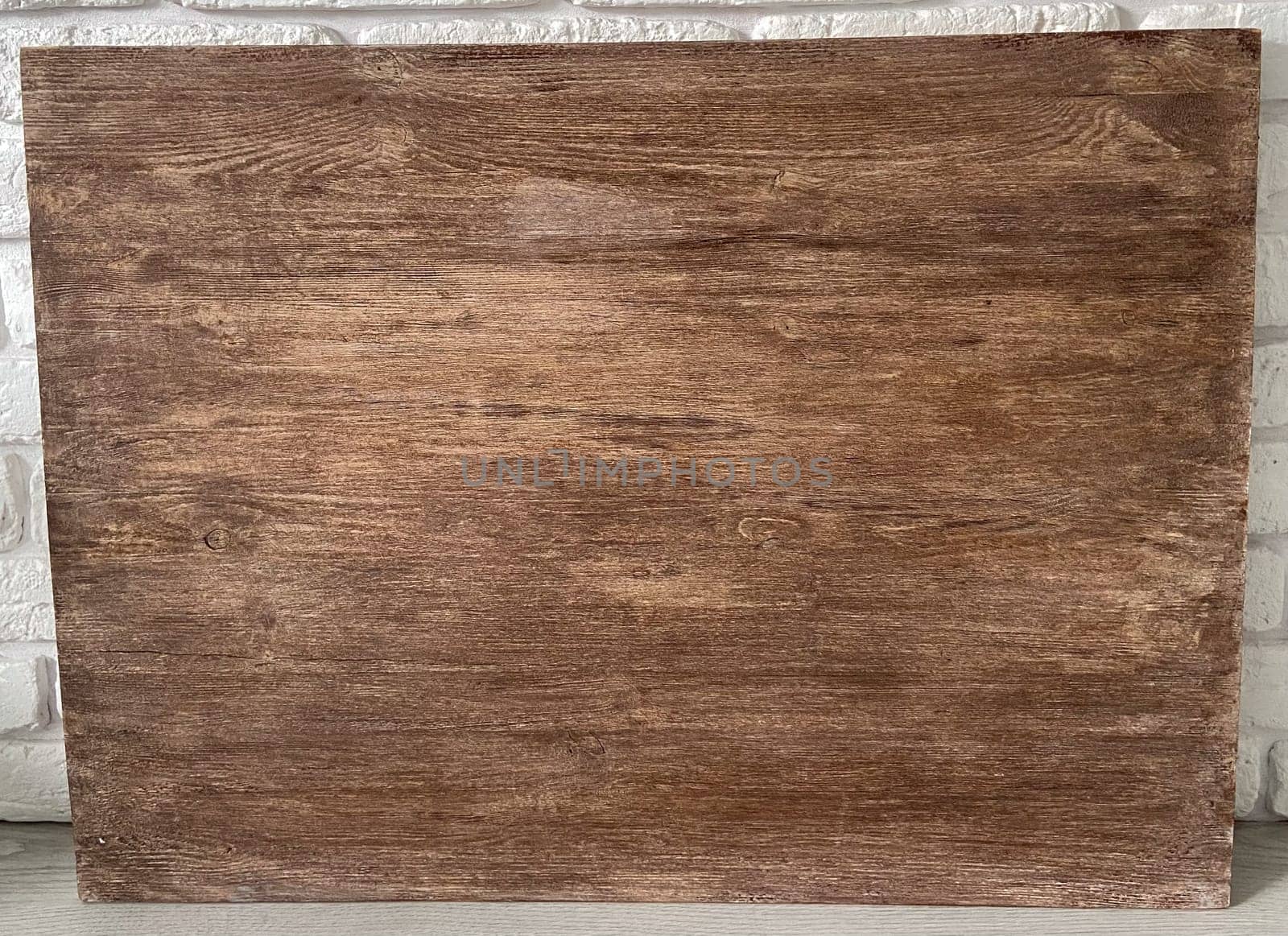Rustic wooden panel leans gracefully against a textured white brick wall, showcasing its rich grain patterns and natural warmth. Contrast between the organic wood and the structured brick provides a harmonious blend of nature and architecture.