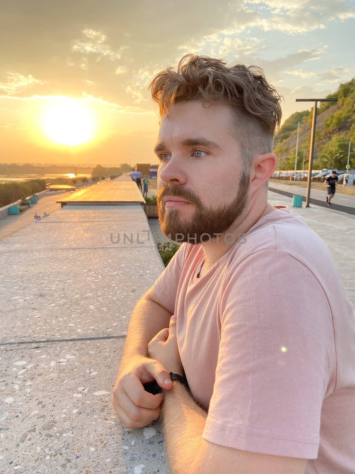 Bearded man gazes thoughtfully at the sunset, leaning against a concrete railing on a scenic promenade. Soft glow of the setting sun creates a serene and reflective atmosphere.