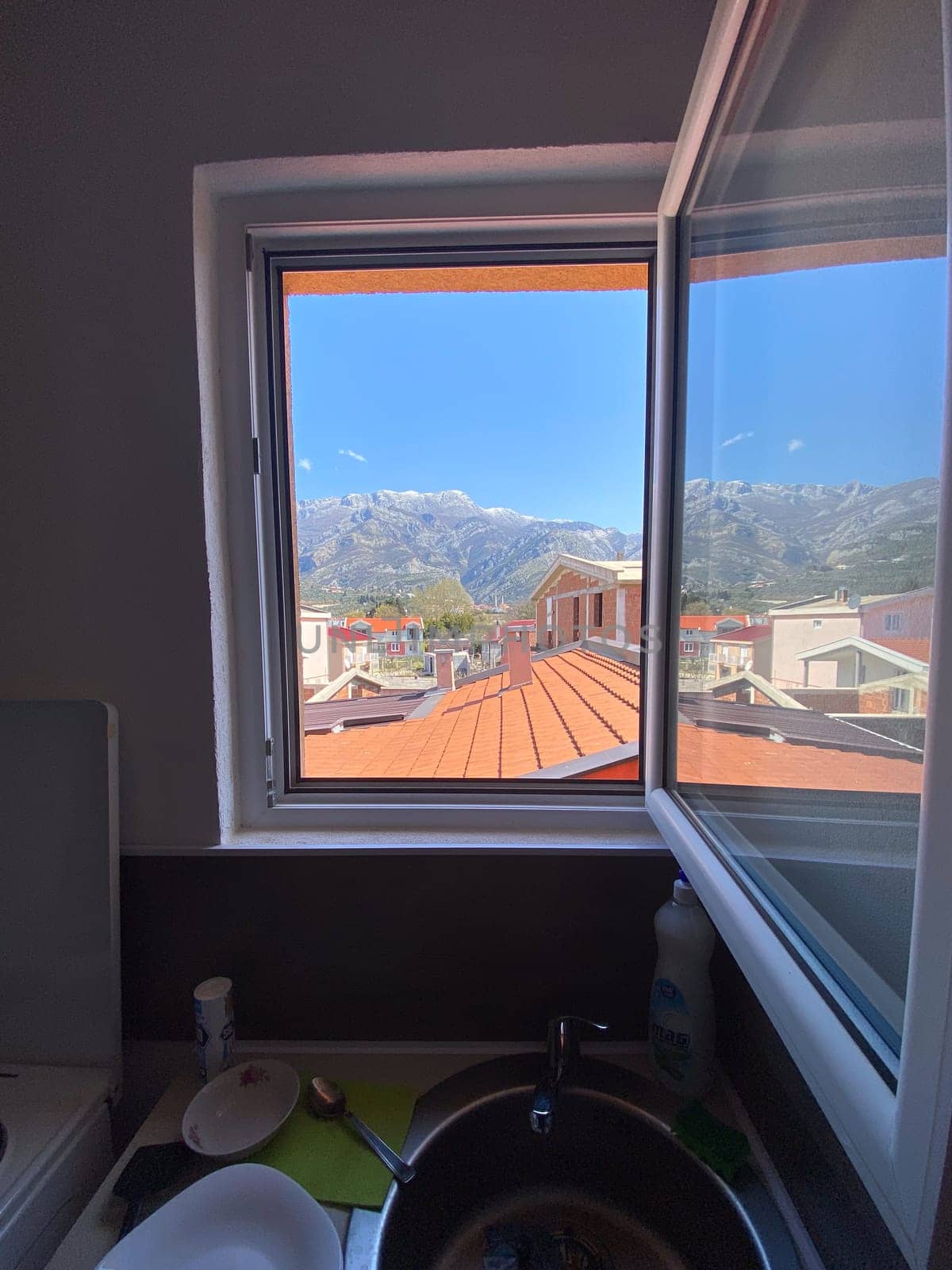 View of distant mountains framed perfectly by an open window, revealing rooftops and a clear blue sky. Indoor sink area contrasts with the vibrant outdoor scenery