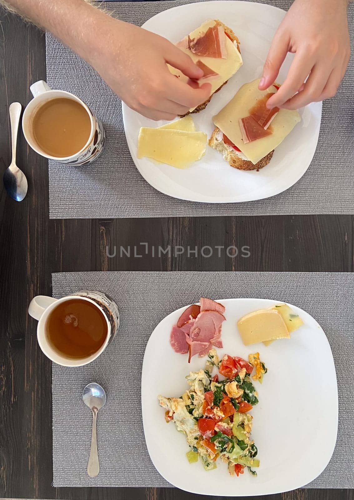Cozy breakfast setting shows a person preparing open faced sandwiches while another plate displays a colorful scramble with coffee cups nearby. Portrays a homey and inviting morning meal.